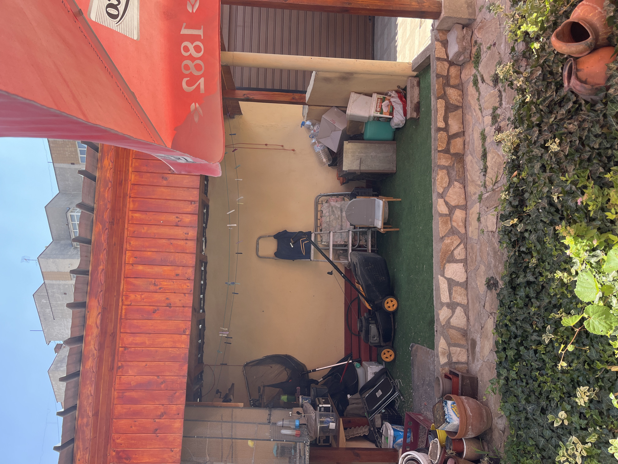 This is an outdoor scene in the city of Pavlikeni, Bulgaria, featuring a covered patio area filled with various garden and storage items. The patio has a wooden slatted roof and walls. On the ground, which appears to be covered with artificial turf, sits a black and yellow lawnmower, a metal folding chair with a light-colored seat, and a small metal cart. To the left, a caged bird is visible, along with shelves holding containers and what appears to be a fishing net. In the background, there are piles of boxes, a large green plastic container, and a ladder with a dark item draped over it. A large red umbrella is partially visible at the top right of the image, casting a shadow. The patio is bordered by a stone wall and a patch of dense green ivy in the foreground. The sky is clear and blue.