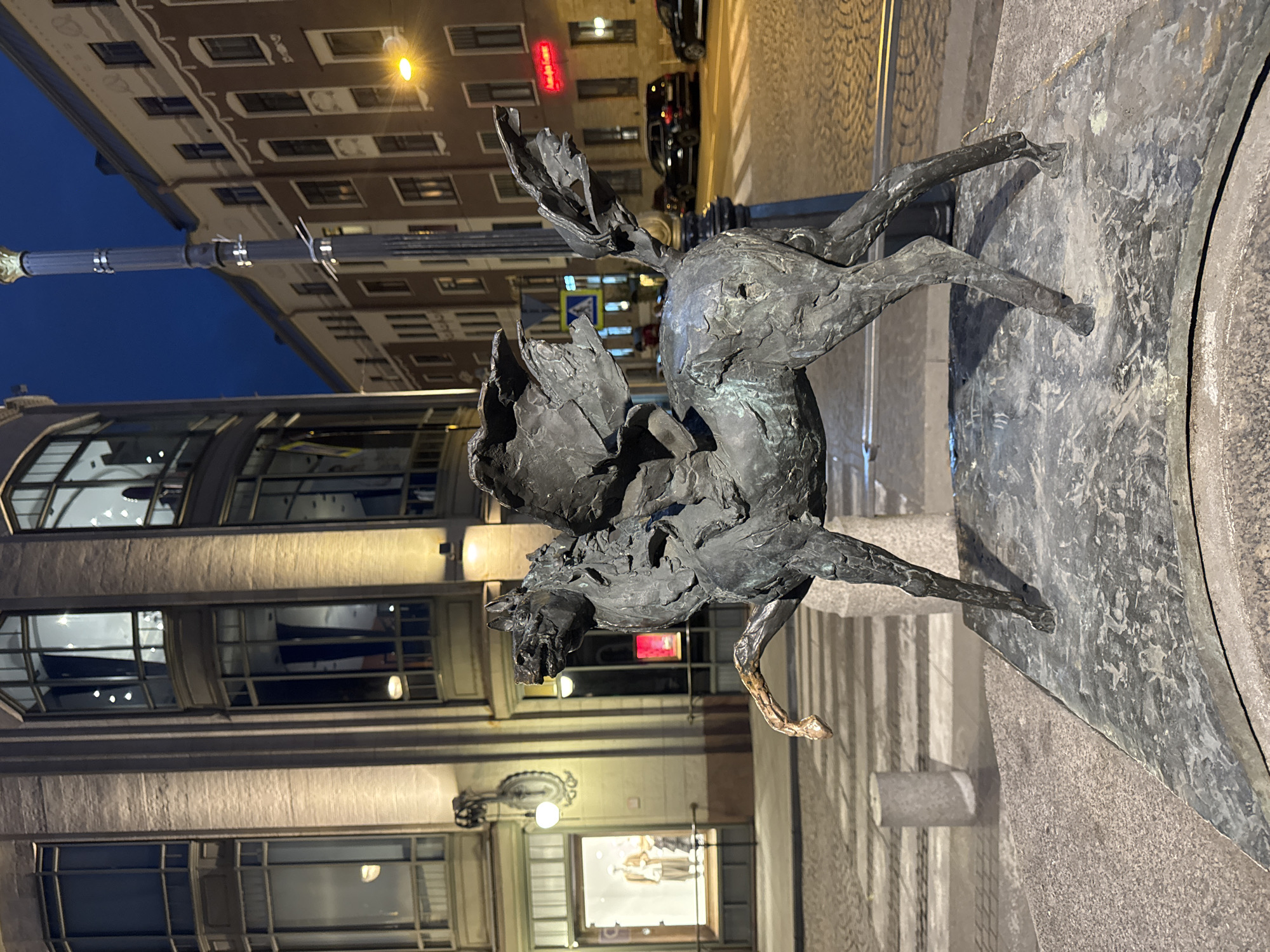 The image shows a bronze statue of Pegasus, the winged horse, in the city of Saint Petersburg, Russia. The statue is positioned on a stone pedestal in front of a building with large windows. The building appears to be a commercial establishment, with mannequins visible in the display windows. Behind the statue, a street is visible, paved with cobblestones. Streetlights illuminate the scene, casting a warm glow. The sky is dark, suggesting it is either dusk or night. Several cars are parked along the street. A lamppost stands to the right of the statue. The overall impression is one of an urban nighttime scene featuring a prominent mythological sculpture.