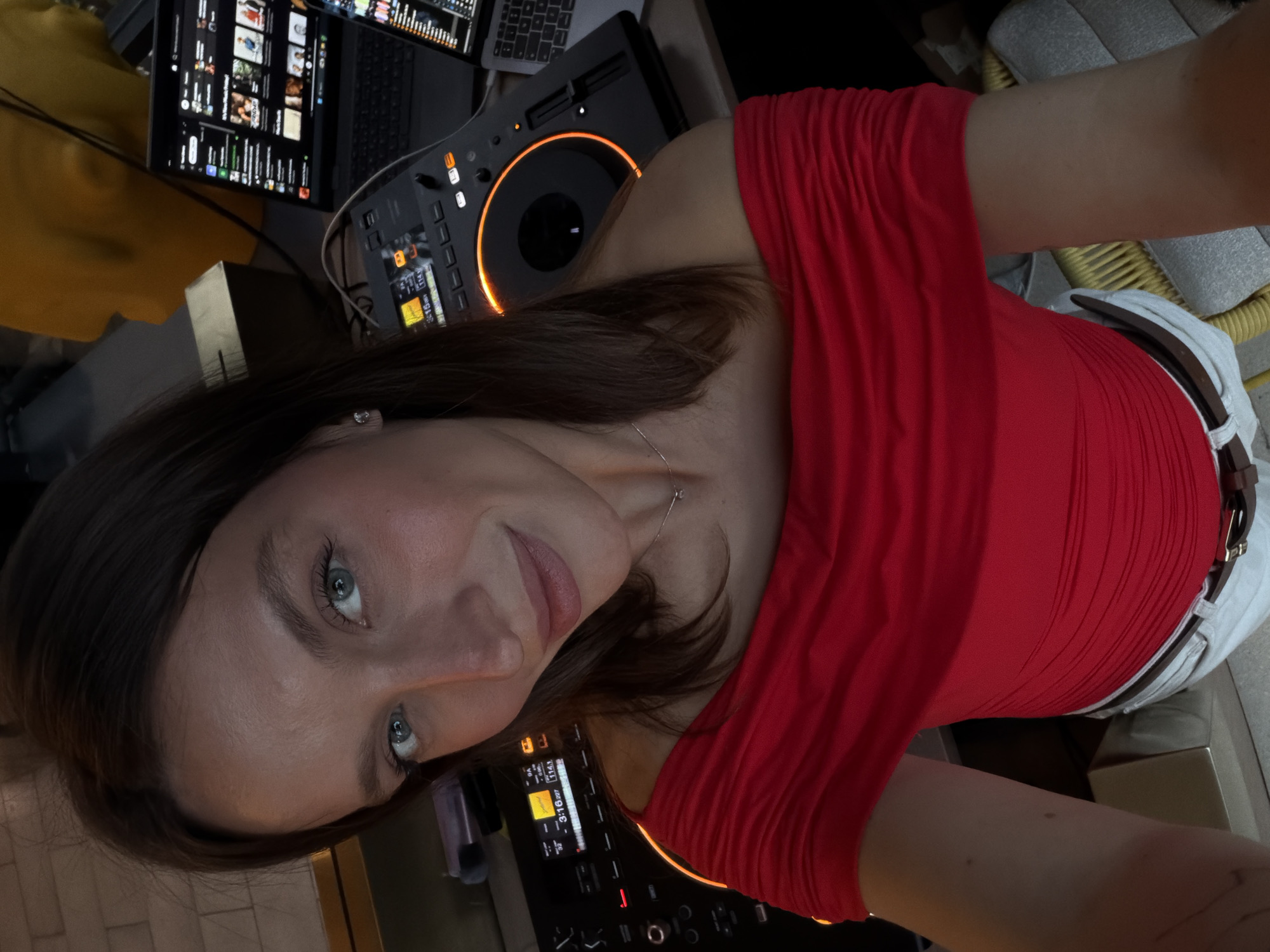 A woman with auburn hair, wearing a red off-the-shoulder top, white pants, and a brown belt, is visible in this selfie-style photograph. She has blue eyes and is looking directly at the camera with a slight smile. The background includes DJ equipment, specifically a digital mixer and computer screens displaying music-related applications. Other objects include a laptop, a tablet, and elements of a sound system. The location is possibly a music venue or DJ booth; no specific landmarks indicate the city of Budva, Montenegro.