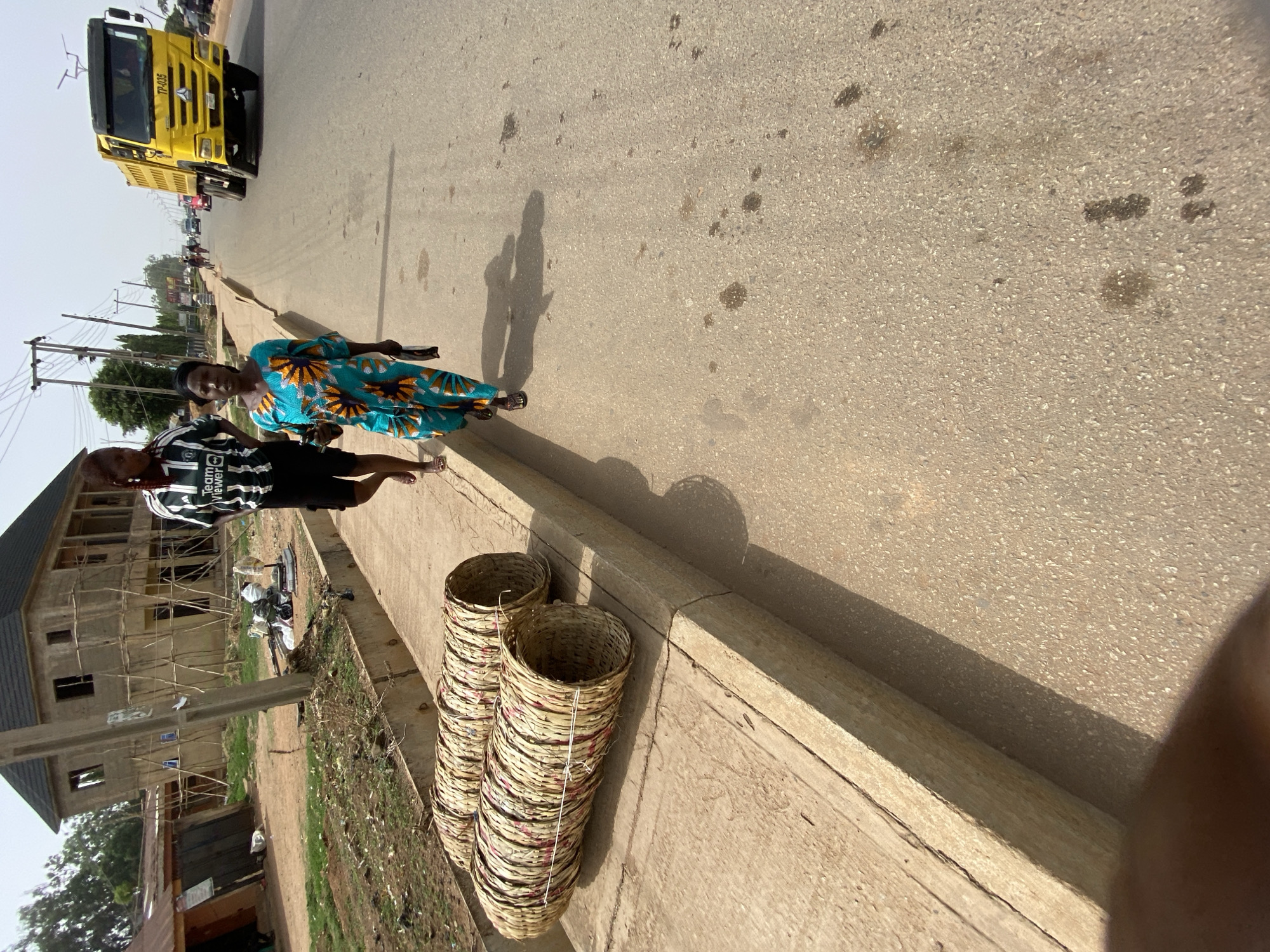 The image captures a roadside scene in what is indicated to be Bukuru, Nigeria, under bright daylight. The sun casts strong shadows, suggesting midday or early afternoon.
In the foreground, on the right, are two people standing on a paved sidewalk or curb. An adult woman, wearing a teal and orange patterned dress, stands closer to the viewer, looking towards the camera. Next to her, slightly behind, is a younger person or child wearing a black and white striped shirt with "TeamViewer" and "Power" visible text (likely a Manchester United jersey) and dark shorts. Their faces appear neutral.
To their left, on the paved road, a large yellow truck is visible, facing away from the camera. The text "TP-035" is legible on its front. Other vehicles can be seen in the distance on the road, suggesting traffic.
Along the right side of the sidewalk, a stack of tall, cylindrical woven baskets is placed. These baskets appear to be made of light-colored natural fibers.
On the left side of the image, behind the people, is an area with rough terrain and some sparse vegetation. Further back, an unfinished multi-story building with wooden scaffolding indicates ongoing construction. Utility poles with overhead lines are also present, extending into the background. The overall scene depicts a bustling, developing urban or semi-urban environment.