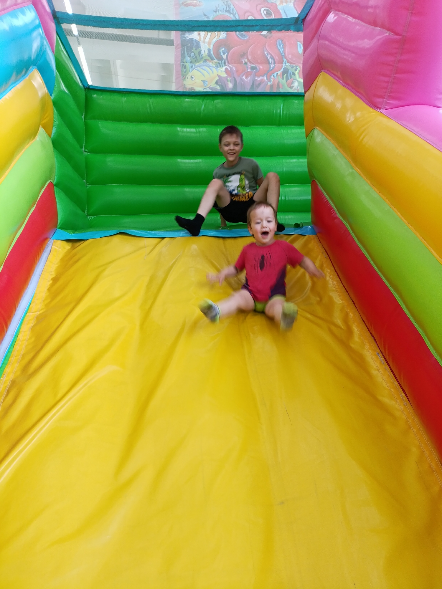 Two boys are on an inflatable slide. One boy is sitting at the top of the slide in a green shirt and black shorts and socks, the other boy is sliding down the slide wearing a red shirt, green shorts, and multi-colored shoes. The inflatable slide is multi-colored with green, yellow, pink, and red sections. A photograph of sea life can be seen behind the inflatable slide, which is located in the city of Chaikovsky, Russia.