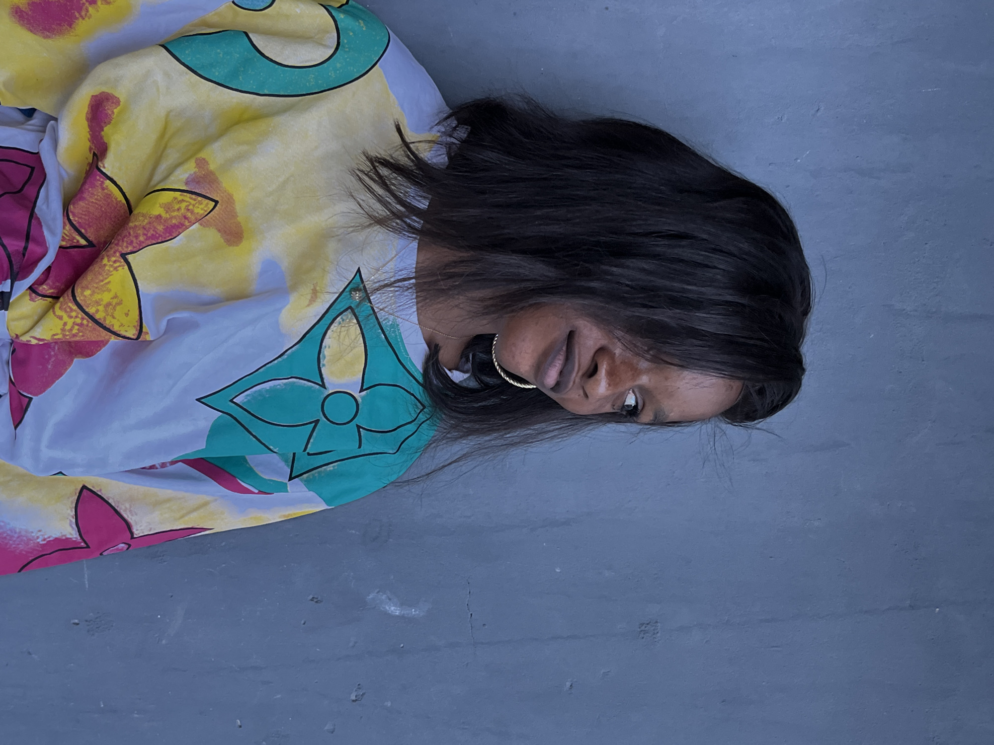 A person with dark hair and skin lies on a blue concrete surface. The person is wearing a light-colored dress with a floral pattern in pink, yellow, and turquoise. The scene is located in Badagry, Nigeria.