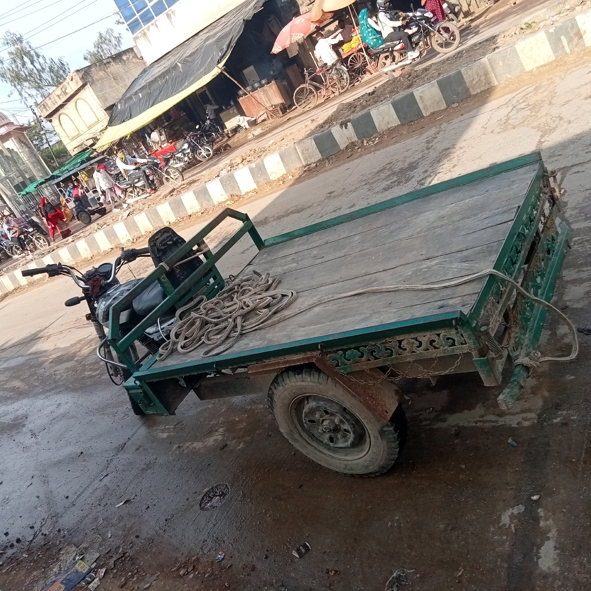 This image depicts a street scene in Phulwara, India. In the foreground, a modified motorcycle with a flatbed trailer is parked on a wet, paved surface. The trailer is constructed with a wooden platform and a green metal frame. A coiled rope lies on the trailer's bed. The motorcycle itself has a dark-colored body and handlebars. In the background, there are buildings with awnings, some appearing to be shops. Several motorcycles and people are visible, suggesting a marketplace or busy street. One person is riding a motorcycle, while others appear to be standing or walking. There is also a bicycle with a basket, and a cart loaded with items. The overall environment appears somewhat cluttered and worn, with debris on the ground. The sky is visible in the upper portion of the image, indicating it is daytime.