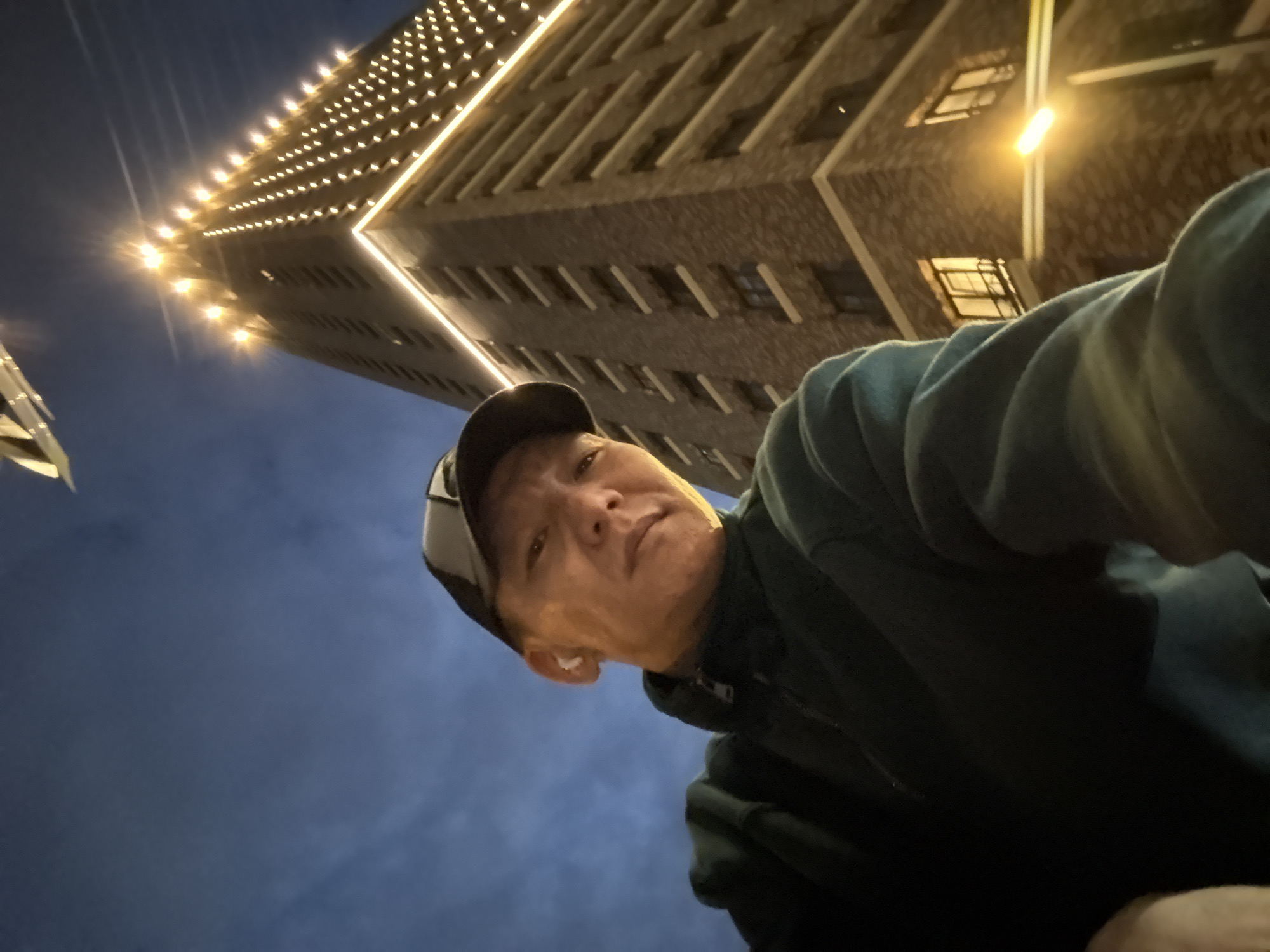A low-angle, wide-shot selfie taken at dusk features a man wearing a dark green hooded jacket and a baseball cap. He has white earbuds in both ears. Behind him, a tall, modern building is illuminated with many small yellow lights along its edges and top, and a brighter yellow light emanates from a streetlamp on the right side of the frame. The sky is a deep blue with some wispy clouds. The man's face is partially illuminated by the building's lights and the streetlamp, giving it a warm glow. The building appears to be residential, with visible windows. The scene suggests an urban environment in Dolgoprudny, Russia, possibly near a new development.
