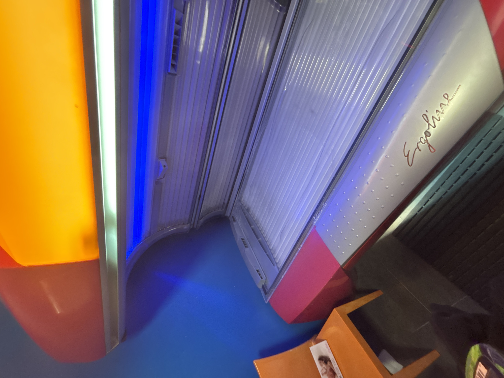 The image displays an interior scene from a tanning salon in Varna, Bulgaria. A horizontal Ergoline tanning bed, featuring a metallic silver-grey base with magenta-red accents, occupies the center-right of the frame. The top canopy of the tanning bed is open, revealing multiple blue fluorescent lamps within the tanning chamber and a white/light green fluorescent tube positioned above it. The upper section of the tanning bed's structure emits an amber-orange glow. To the left, an unoccupied, bright orange chair is positioned against a vibrant blue wall. A white magazine or flyer, partially displaying text "M FOR YOU" and an image of a person, rests on the chair's seat. The floor, visible at the bottom of the frame, is dark in color. No individuals are present, and no activity is depicted.