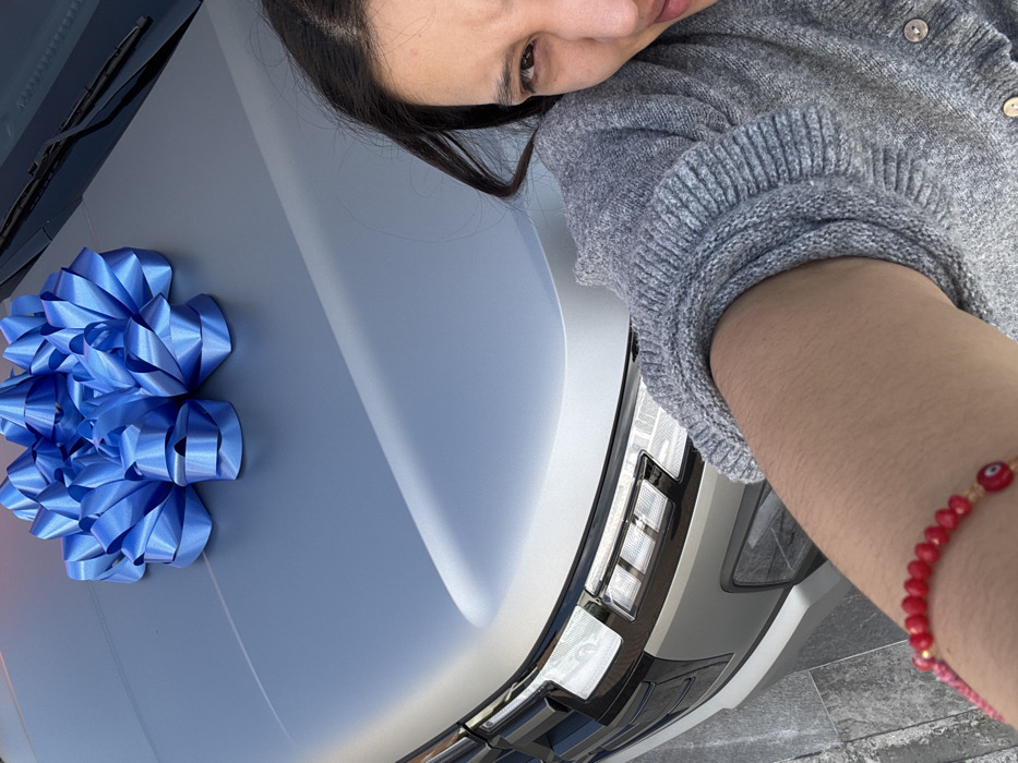 A woman in a grey sweater leans against the hood of a silver car. A large blue bow sits on the hood near the windshield. The car's headlight is visible on the right side. The woman's arm extends into the frame on the right, featuring a red beaded bracelet. The image is angled, presenting a close-up of the woman and the car.