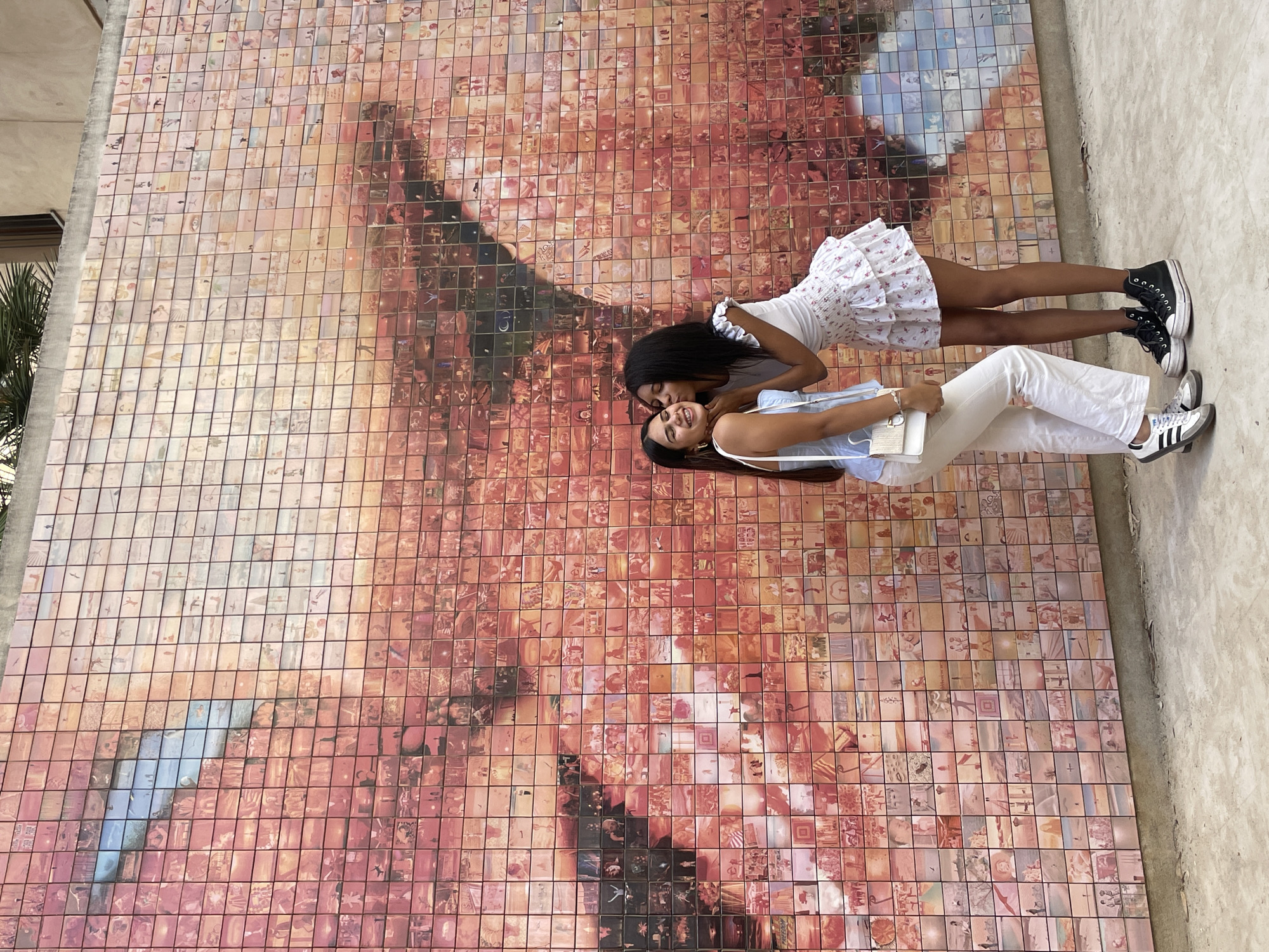 Two young women are posing in an outdoor setting in front of "El Beso" (The Kiss), a large photographic mosaic mural by Joan Fontcuberta, located in Plaça d'Isidre Nonell, Barcelona, Spain. The mural is composed of thousands of individual square tiles, each containing a smaller photographic image, which collectively form the larger image of two lips kissing, predominantly in shades of red, orange, and pink with blue accents.
The woman on the left, with long dark hair, wears a light blue sleeveless top, white high-waisted trousers, white sneakers with grey accents, and carries a white handbag with a gold chain strap. She is smiling with closed eyes, her head tilted to the right, receiving a kiss on her left cheek. The woman on the right, with long dark curly hair, wears a white top with ruffled sleeves, a white mini-skirt with a subtle floral pattern, and black high-top sneakers with white soles. She is leaning in, her lips touching the cheek of the other woman.
The ground in the foreground is a light grey concrete pavement. Above the mosaic, a concrete ledge and part of a building facade with green foliage are visible. The scene is illuminated by natural daylight.