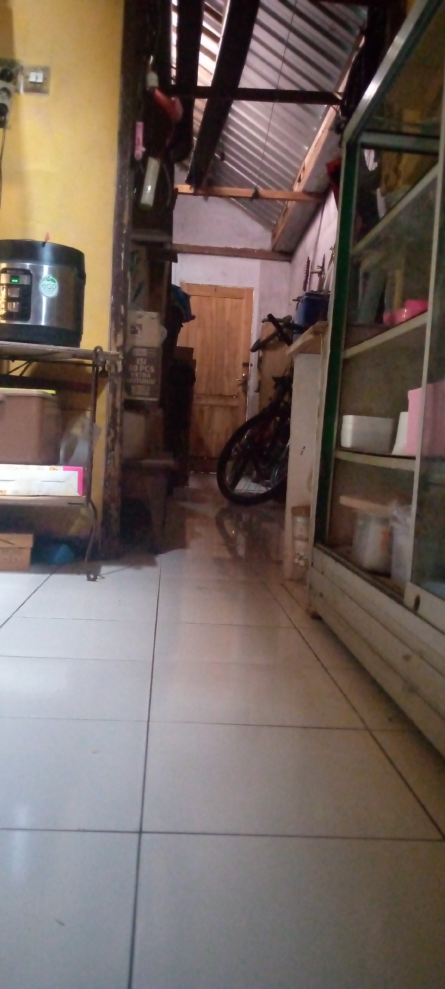 The image depicts an interior scene, possibly a home or shop, in the city of Sumberwono, Indonesia. The foreground is dominated by light-colored tiled flooring. To the left, a silver rice cooker with an "eco" logo is placed on a metal shelf. A wooden door is visible in the background, to the right of a dark-colored vertical support. Further to the right, a bicycle with black tires is parked. A glass display cabinet, likely containing various items, occupies the far right side of the image. The ceiling appears to be constructed with corrugated metal sheets supported by wooden beams. The overall environment suggests a functional and somewhat cluttered space.