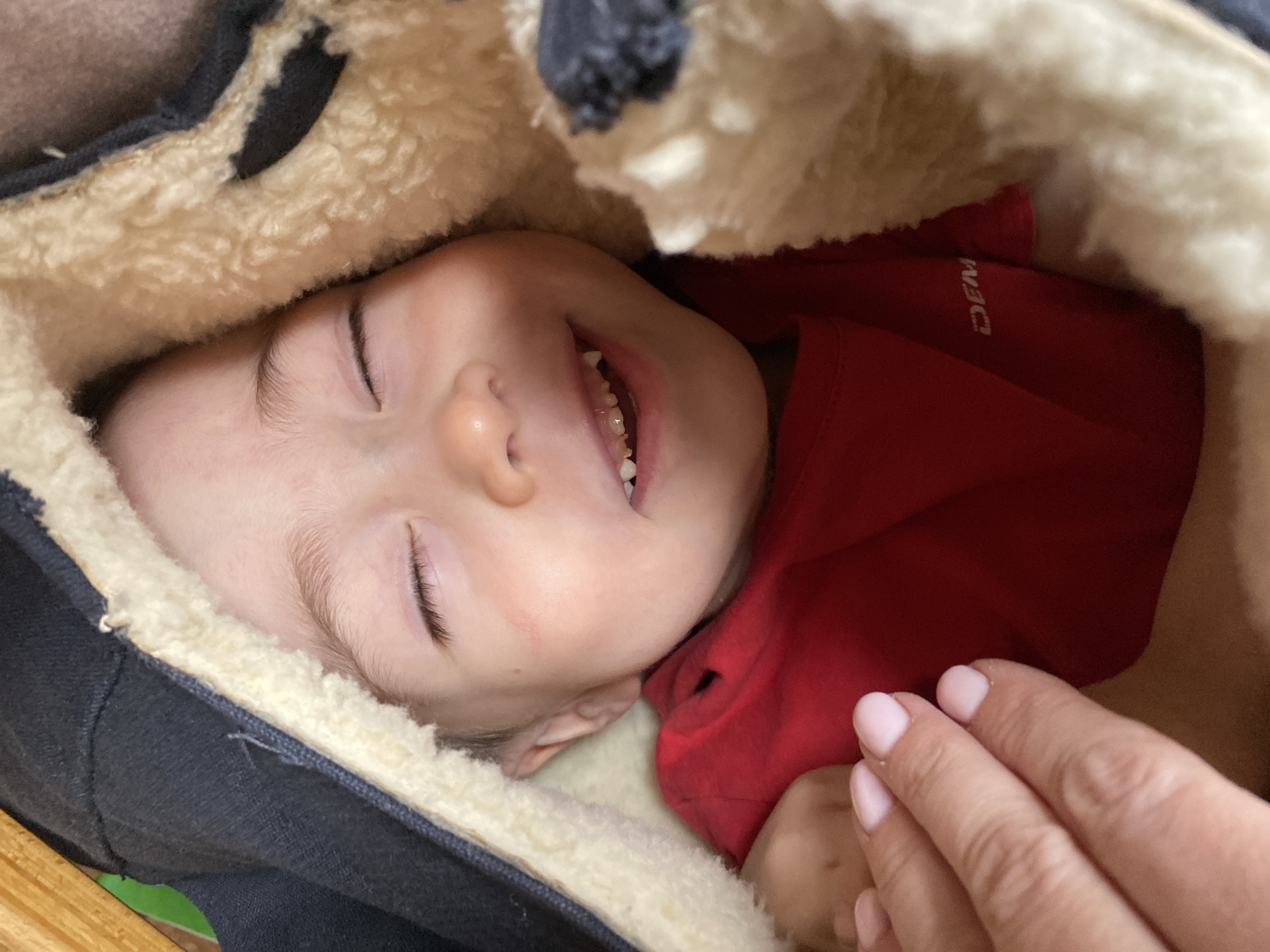 A close-up image shows a baby nestled inside what appears to be a dark blue and beige fuzzy-lined carrier or hooded coat. The baby, wearing a red shirt, is lying on its side with its eyes closed and a slight smile, revealing some teeth. A hand with painted nails is visible near the baby's torso. The location is not discernible from the image, so it is not possible to confirm it is in Ольховка, Russia.