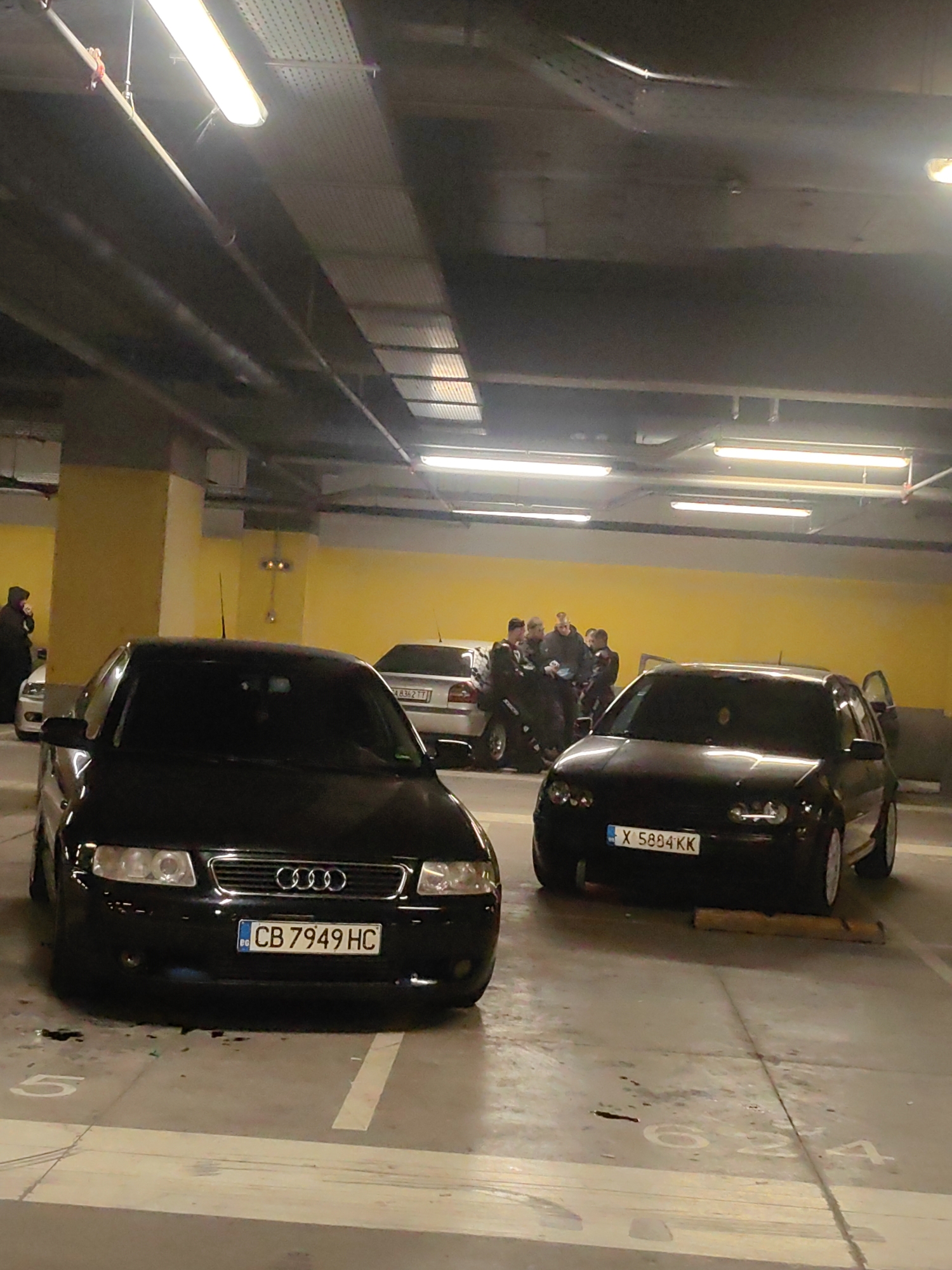 The photograph depicts an indoor parking garage in Sofia, Bulgaria. Two black cars are parked in the foreground, an Audi A3 with license plate CB 7949 HC and a Volkswagen Golf IV with license plate X 5884 KK. In the background, a group of approximately seven individuals are gathered around a silver car, seemingly engaged in conversation. The garage is illuminated by fluorescent lights overhead, and parking space numbers are visible on the floor.