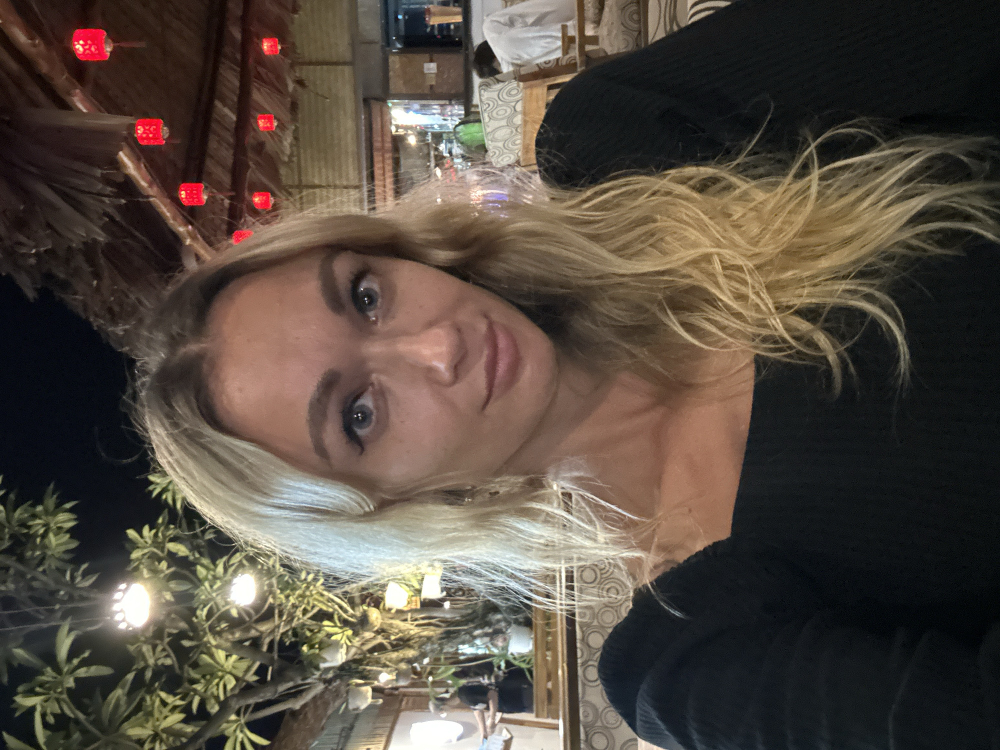 A medium shot, outdoor, selfie-style image shows a fair-skinned woman with blonde, wavy hair and blue eyes. She is wearing a black top. The woman is positioned in the foreground, with her face taking up the majority of the frame. In the background, there is a rustic, open-air seating area with thatched roofing. Red lanterns are hanging from the roof. Trees with green foliage and hanging lights are visible on the left side of the frame. The scene appears to be at night.