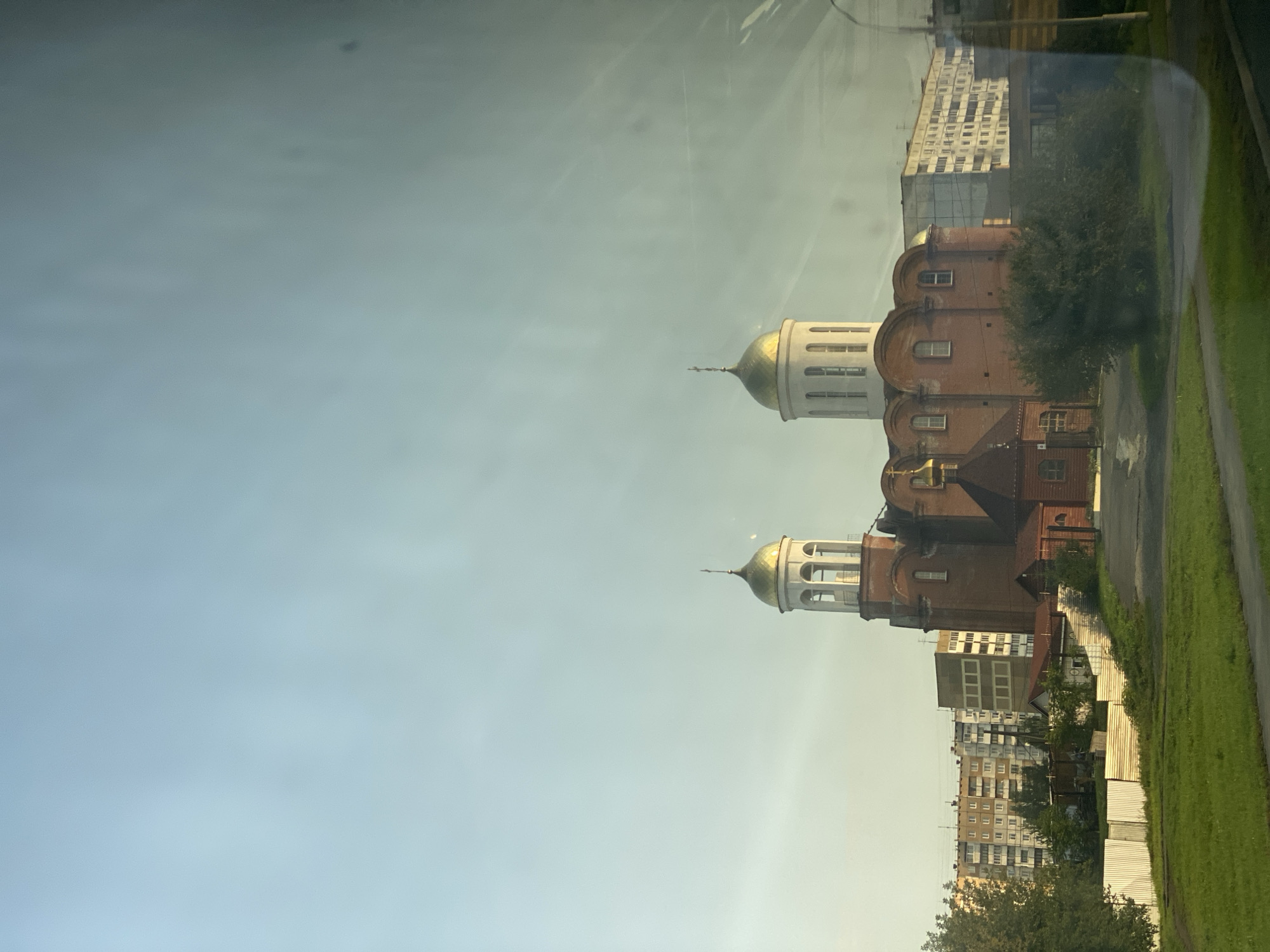 The image captures an Orthodox church with two golden domes, likely in the city of Kemerovo, Russia. The church is constructed of red brick and features a bell tower on the left. To the left of the church, there are several residential buildings, some appearing to be apartment blocks, and a white metal fence. The foreground shows a grassy area and a paved path. The sky is clear and light blue, with some light flares visible, suggesting the photo was taken through a window. The overall scene depicts a juxtaposition of religious architecture with urban residential structures under a bright sky.