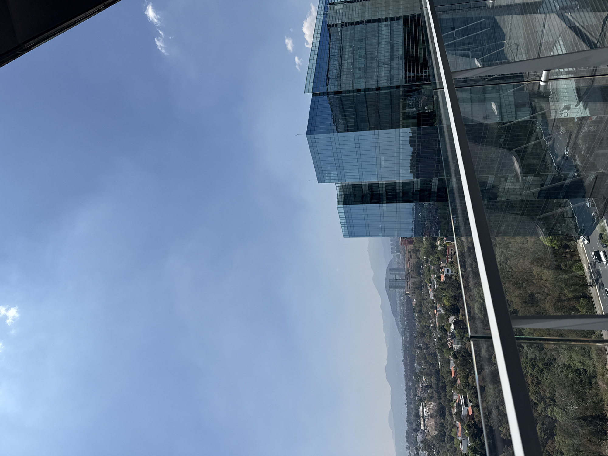 The image shows a view from a high vantage point, likely a tall building in Mexico City, Mexico. The sky is a clear blue with some haze and few scattered clouds. The cityscape below includes buildings, trees, and roads. There's a cluster of modern, glass-facade skyscrapers on the right side of the image. In the background, a mountain range is partially obscured by haze. A road with cars is visible between trees. The composition is angled, adding a sense of height and perspective to the scene.