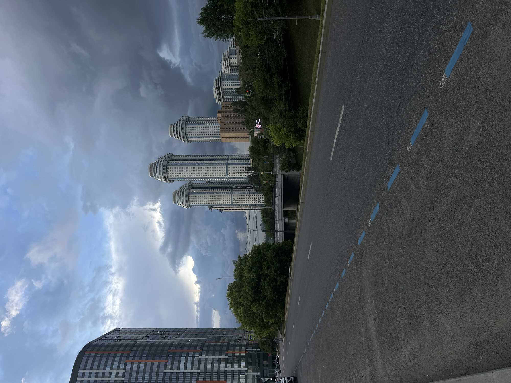 The image captures a street scene in Moscow, Russia, likely on a cloudy day with dramatic storm clouds gathering in the sky. The foreground features an asphalt road with dashed blue markings, indicating a traffic lane or parking area. To the left, a tall, modern building with a curved facade, featuring a pattern of dark and orange vertical accents, dominates the skyline. In the mid-ground, several distinctive, cylindrical white high-rise buildings with rounded tops stand clustered together, characteristic of some residential complexes in Moscow. Adjacent to these, a dense cluster of green trees forms a lush barrier. A concrete bridge crosses a waterway or depression in the distance, with traffic lights visible. The overall impression is one of an urban environment with a mix of contemporary and established architecture, under a moody, atmospheric sky.