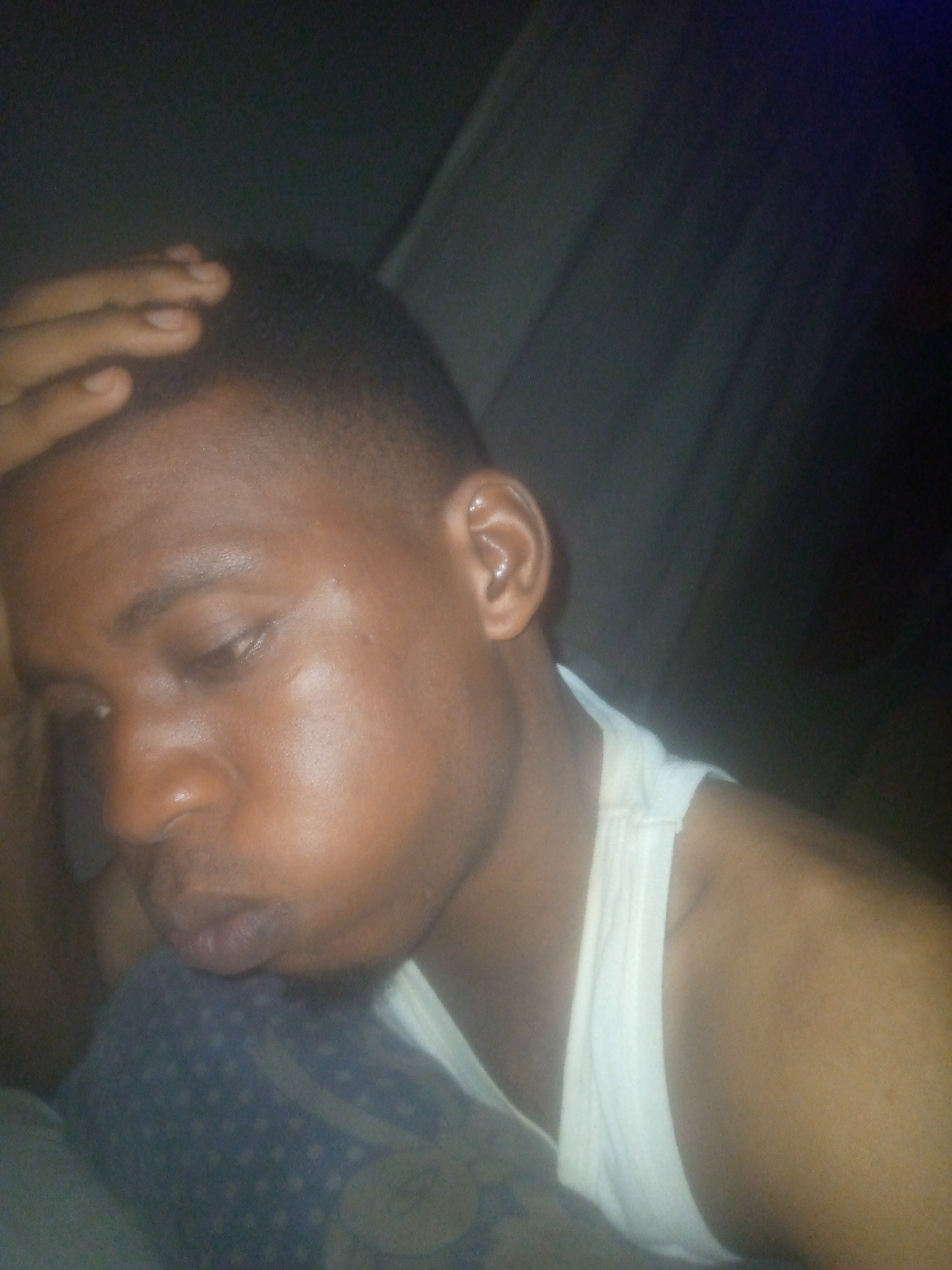 A close-up shot depicts a person with dark skin and short, dark hair. The individual has one hand resting on their head. The person is wearing a white tank top. The background appears dark and indistinct. The location is Unknown, Unknown.