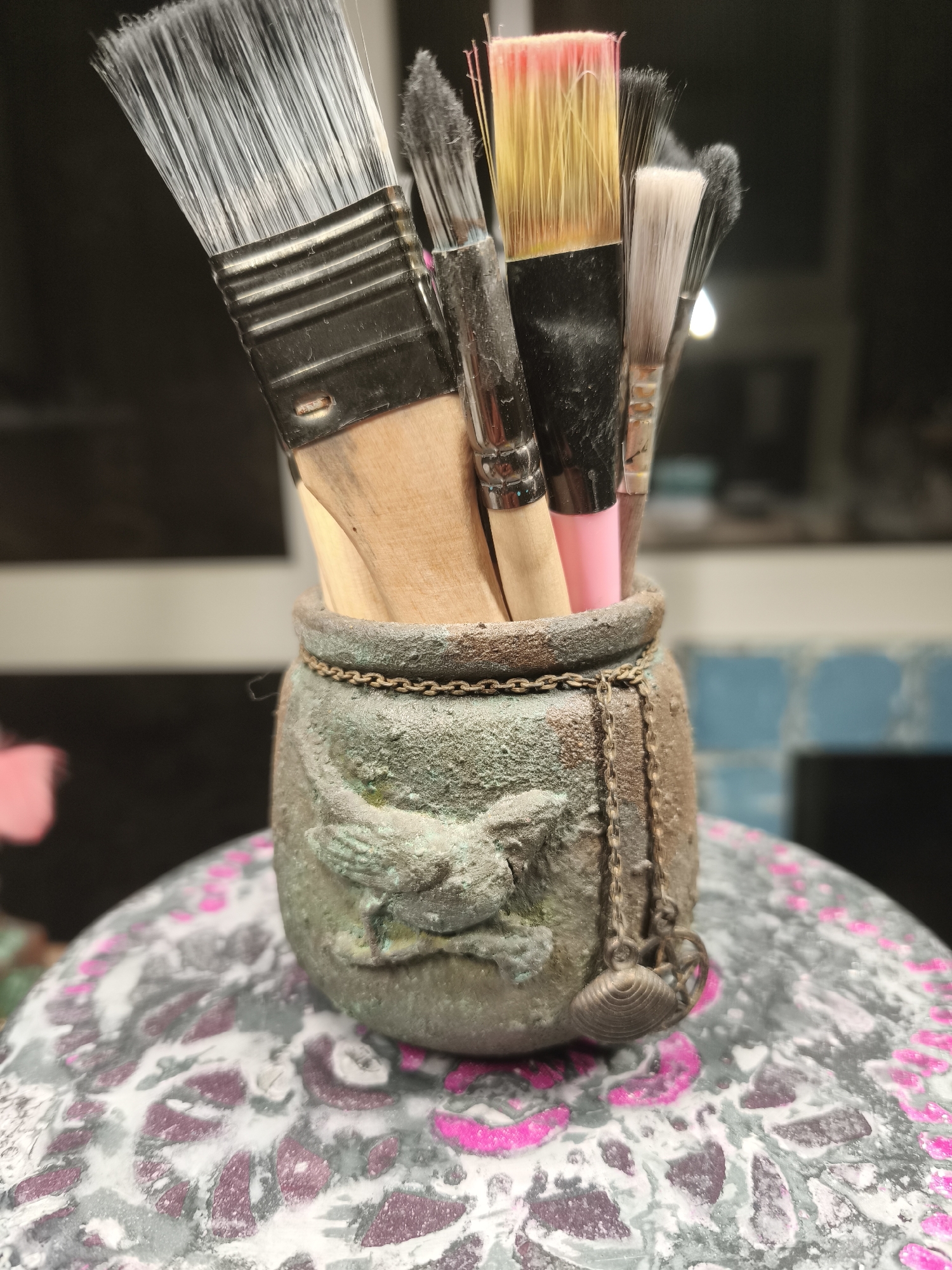 A close-up, low-angle shot focuses on a collection of paintbrushes held upright in an ornate, weathered green pot. The pot has a textured surface with raised designs, including what appears to be a bird motif, and is adorned with a delicate chain and a small shell charm. The bristles of the paintbrushes, varying in size and color, are prominently displayed at the top. Some bristles are white, others are a mix of yellow and pink, and some are black. The wooden handles of the brushes are visible, along with their metal ferrules. The pot rests on a round, textured surface with a pattern of white and dark gray marbling interspersed with bright pink accents. The background is out of focus, showing dark and light horizontal bands, suggesting an indoor setting with a window or shelf. The scene is illuminated from the front, casting soft shadows. There are no people visible in the image, and no explicit details place the scene in Камышеваха, Russia.
