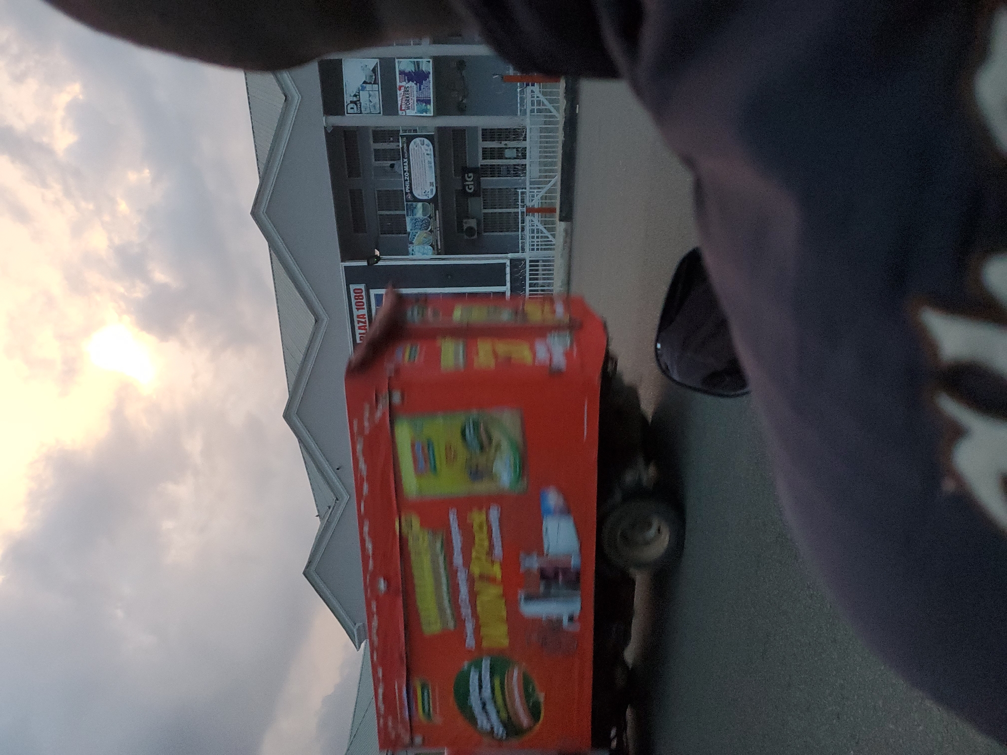 The image shows a blurred view from a vehicle in Jos, Nigeria. A red delivery truck with branding is visible on the left side of the frame, parked on the side of a road. In the background, a modern building complex with a white roof and grey facade is present. Signage on the building indicates "PLAZA 1080". The sky is overcast with the sun peeking through the clouds. The foreground is dominated by the blurred shoulder and head of the photographer. The overall scene suggests an urban environment during daytime.