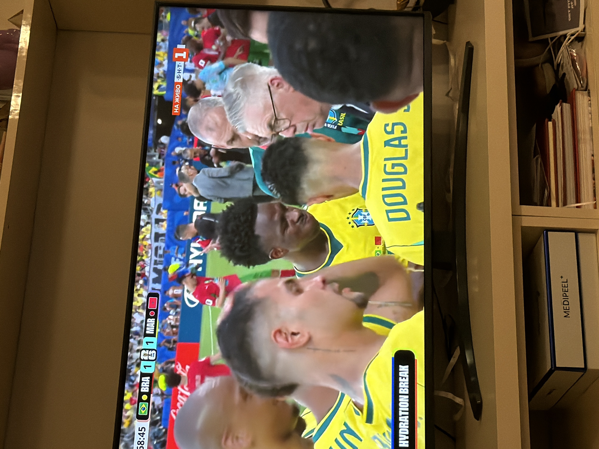 The image shows a television screen displaying a soccer match. The score is 1-1 between BRA and MAR, with the game in the 68th minute and 45 seconds. The text "HYDRATION BREAK" is visible in the lower left corner of the screen. Several people are visible in the foreground, appearing to be watching the television. One person in a yellow soccer jersey with "DOUGLAS" printed on the back is prominent. The television is placed on a shelf, and below the shelf, a box labeled "MEDIPEEL +" is visible, along with stacks of books and papers. The overall setting suggests a domestic environment. The information about the city of Sofia, Bulgaria, is not ascertainable from the visual content of the image itself.