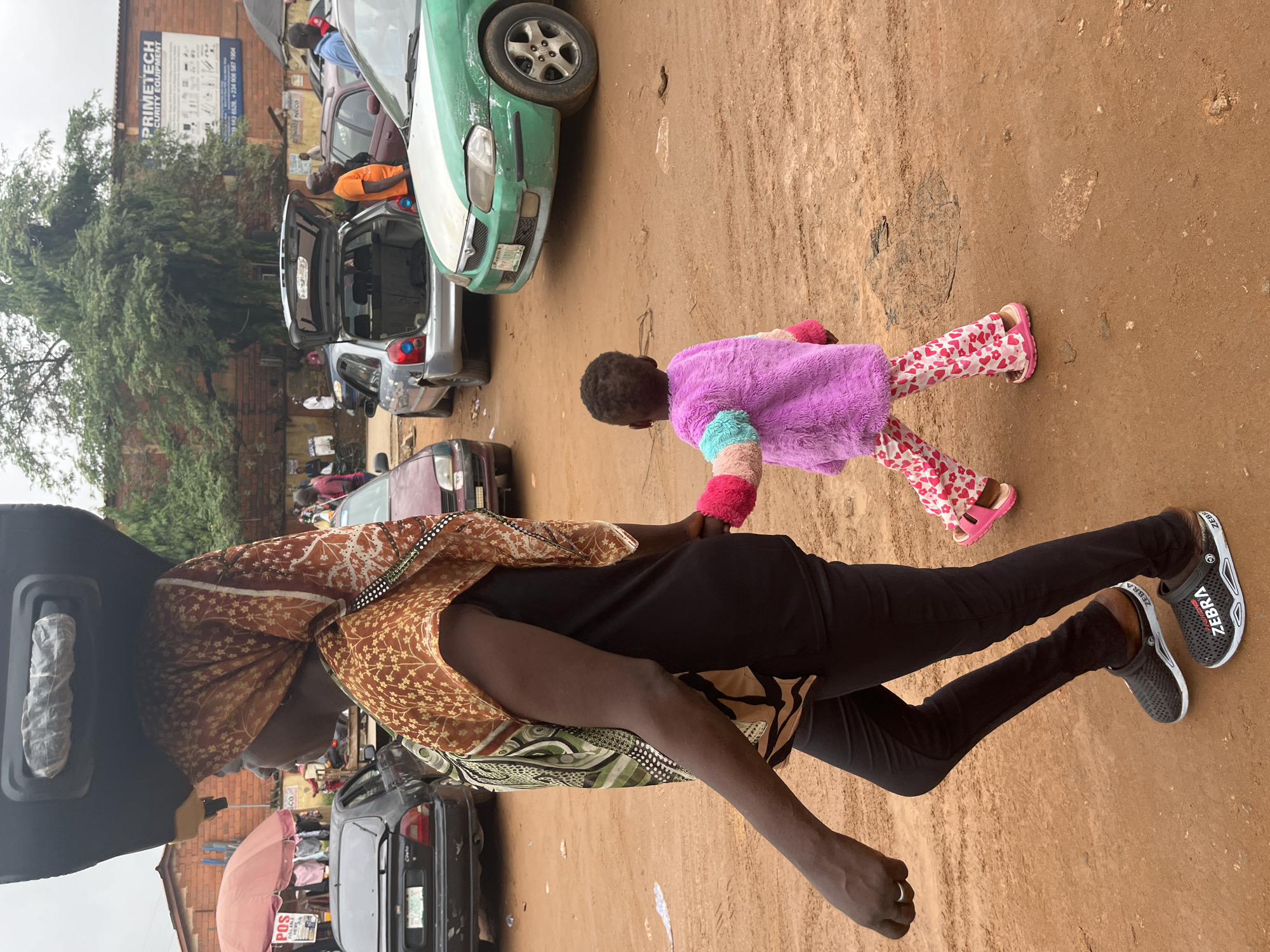 In a dusty, outdoor market area in Abuja, Nigeria, a woman is walking to the left, carrying a large black object on her head. She is wearing a black top, black pants, and patterned headscarf. Her arm is extended to the left, and she is wearing a ring on her left ring finger. To her right, a small child is walking in the same direction, holding her hand. The child is wearing a purple furry jacket and pink pants with a heart pattern, and pink sandals. Several vehicles are parked in the background, including a green and white sedan, a silver hatchback with its trunk open, and a dark sedan. A brick building with signage is visible behind the vehicles. The ground is dirt and appears to be uneven with some puddles.