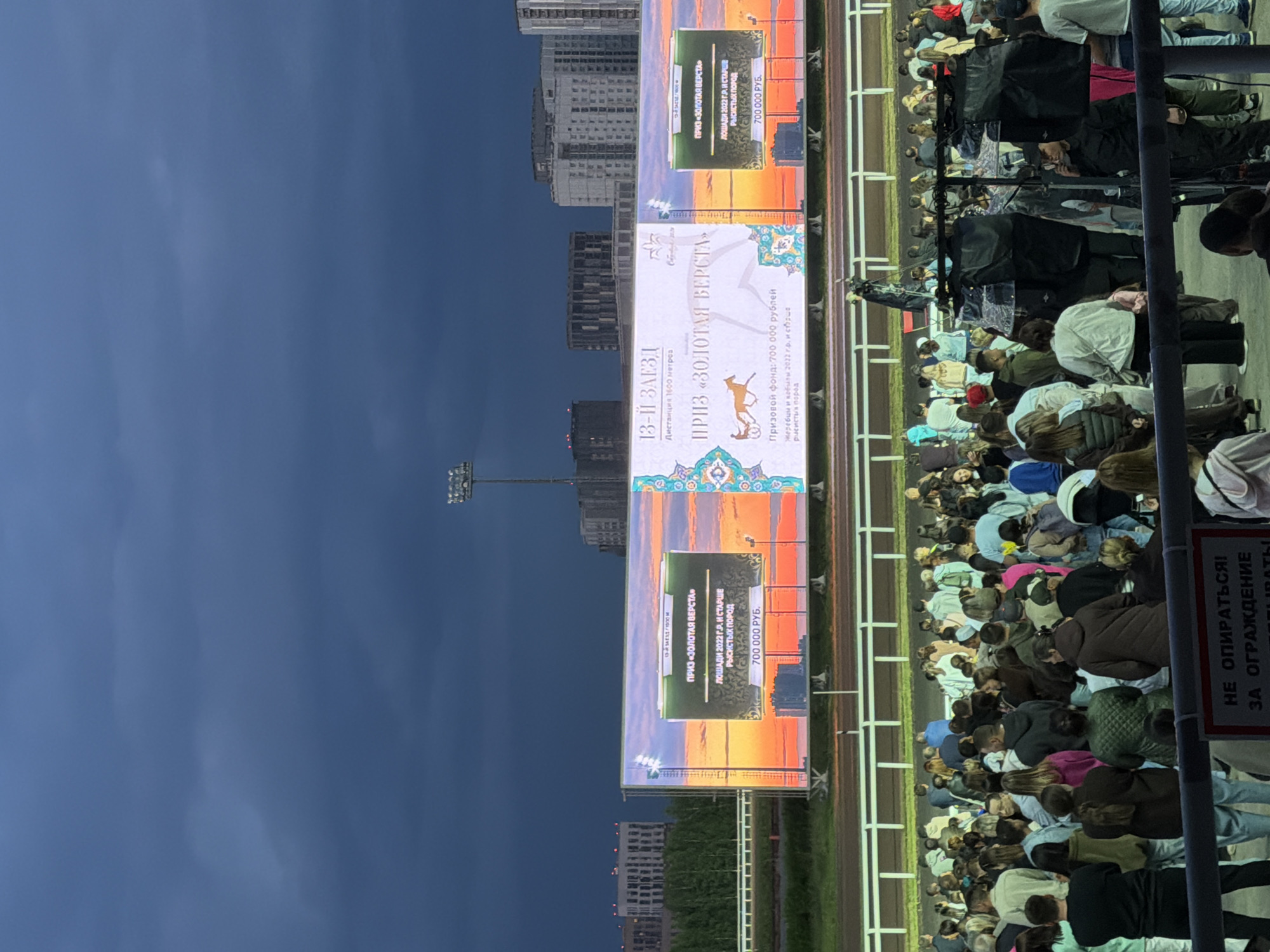 The image depicts a scene at a horse racing track under a dark, cloudy sky. A large LED screen displays information about an upcoming race, showing "PRIZE 'GOLDEN VERST'" and details about prize money and horse breeds. A crowd of spectators is gathered, some standing and some seated, looking towards the screen or the track. In the background, modern buildings and trees are visible. The overall atmosphere suggests an outdoor event taking place in the evening.