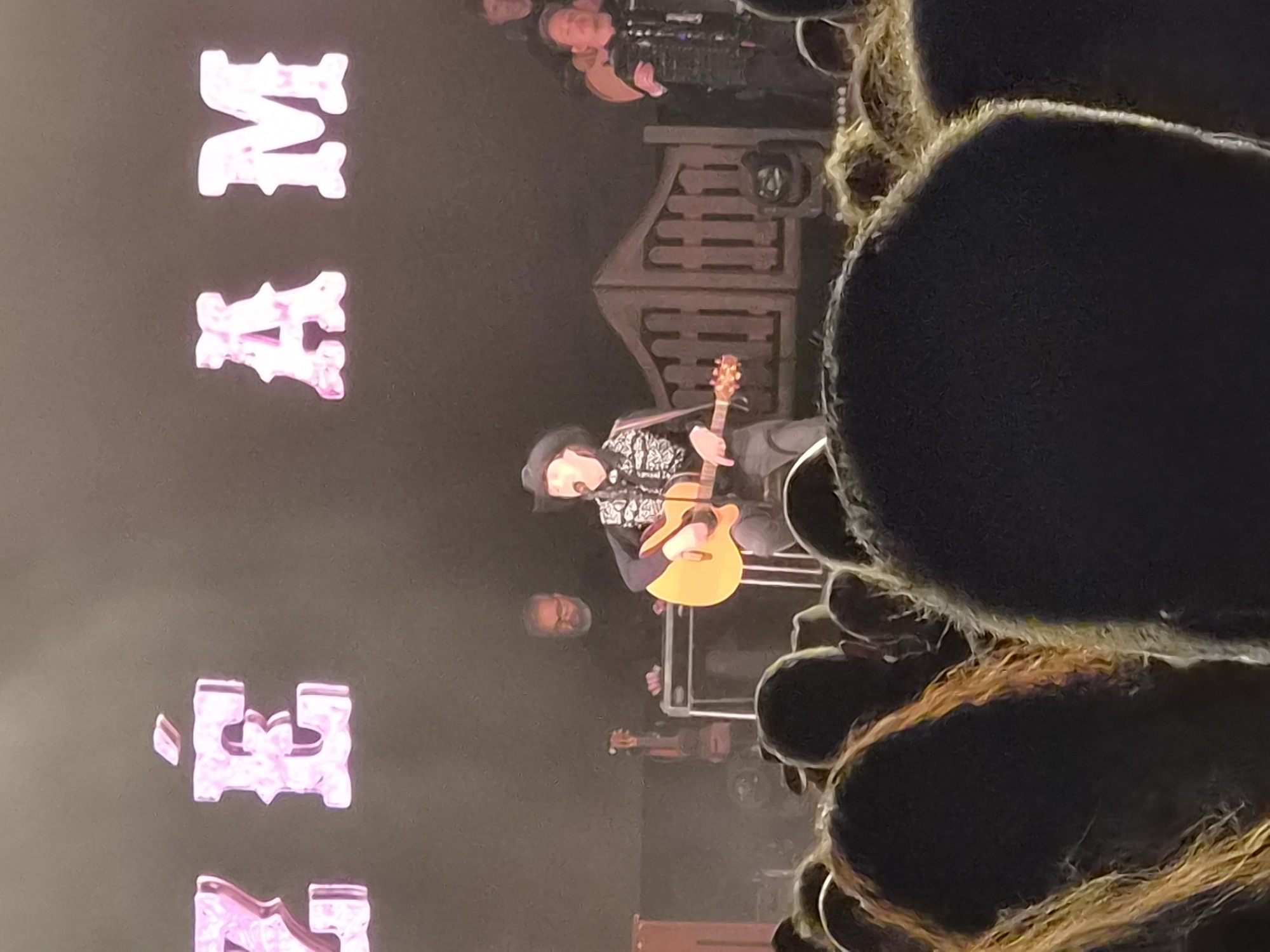 The image shows a musician playing an acoustic guitar on a stage. He is wearing a dark hat and a patterned shirt. Behind him is a backdrop that appears to be a wooden structure. To the left of the musician, there are large pink letters that spell out "MAZE". In the foreground, there are blurred figures, possibly part of the audience. Other people are visible on stage with the musician. The photograph was taken at the Mares Vivas festival in Barcelos, Portugal.