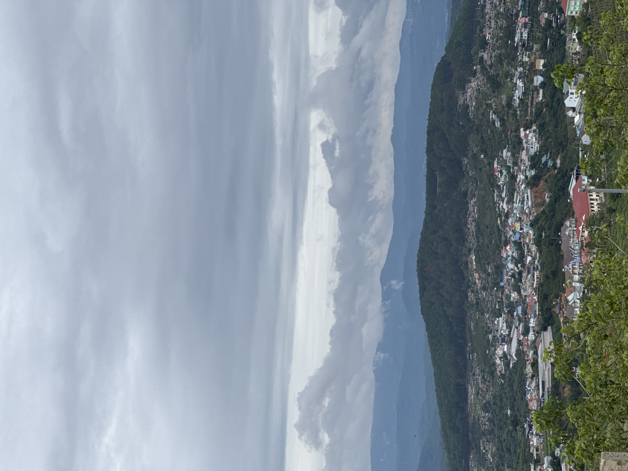 The image depicts a panoramic view of Trường Thọ, Vietnam, on an overcast day. The left portion of the frame is dominated by a gray, cloudy sky, transitioning to a thinner, brighter band of clouds closer to the center. In the middle ground, a series of forested mountains gradually fade into the distance with a blueish hue. The right side of the image shows a dense collection of buildings, likely homes and businesses, clustered along the slopes of a tree-covered hill. The buildings are of various colors and sizes, with red roofs visible among them. A cluster of green foliage occupies the extreme right edge of the frame, obscuring a portion of the urban landscape.