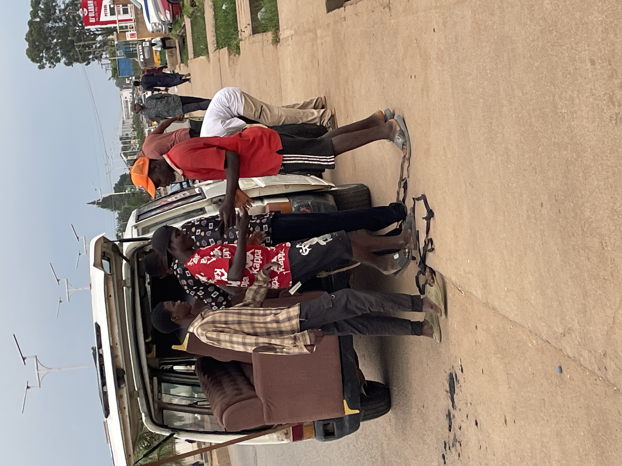 In Barakin Lerawi, Nigeria, on a cloudy day, a group of individuals is engaged in the process of loading a sofa into the back of a white van. The van's rear hatch is open, revealing the interior where the brown sofa is positioned. Several young men are visible, some appearing to be directly involved with the sofa and the van, while others stand nearby.
One man in an orange cap and a red t-shirt is interacting with another individual. Another man, wearing a plaid shirt and dark pants, stands to the left of the van. A third man, in a red t-shirt with "Kappa" written on it, is also present, as are other figures in the background. Straps are visible on the ground near the van, suggesting they are being used or have been used to secure the sofa.
The scene takes place on a paved surface, likely a roadside, with a curb and grassy area to the right. In the background, a busy street with vehicles and buildings can be seen, including a sign that reads "AF'OLABAN PETROLEUM." Streetlights are also visible in the distance. The overall atmosphere appears to be one of a typical urban street activity.