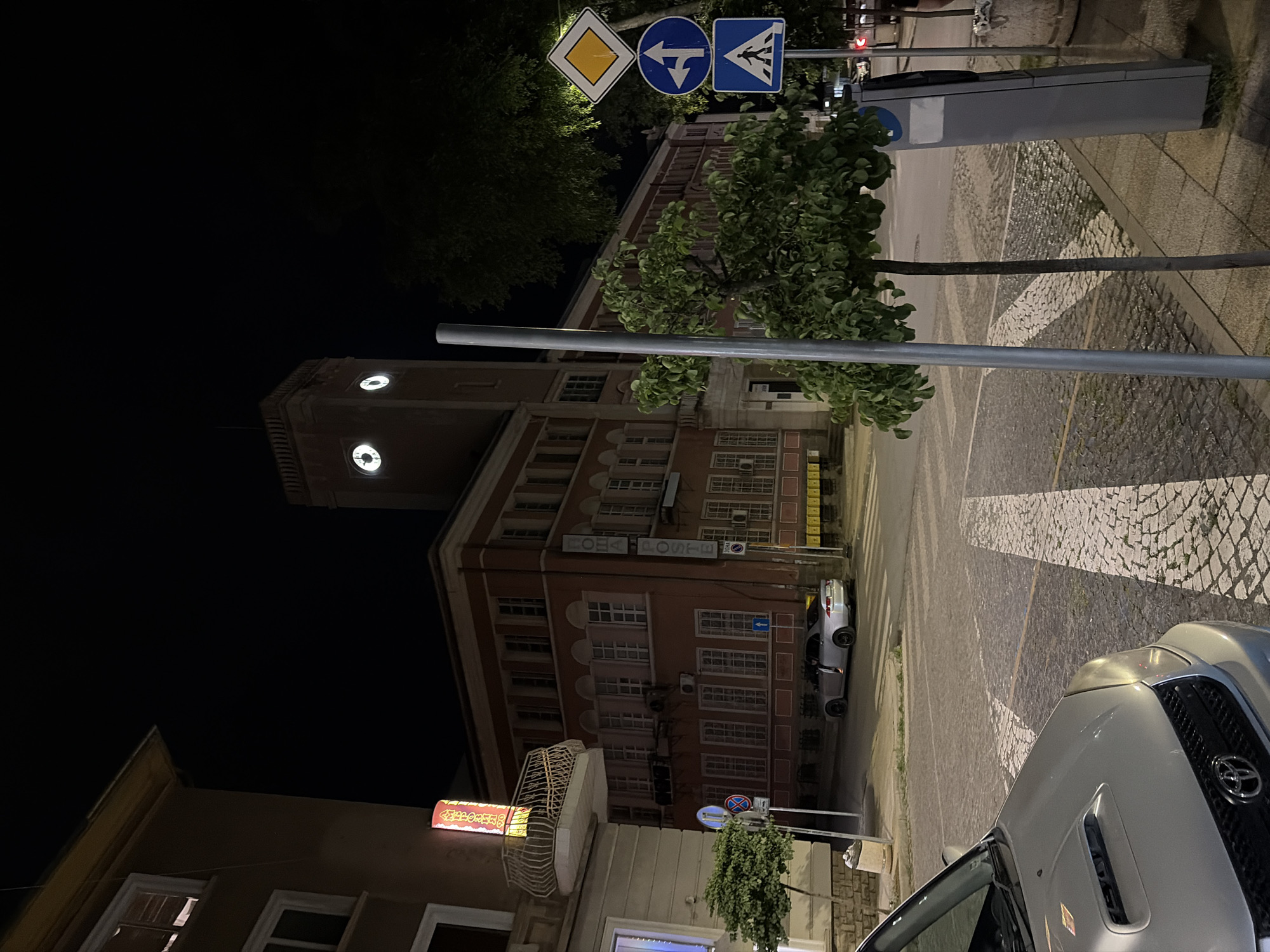 This is a nighttime photograph taken in Stara Zagora, Bulgaria. The scene captures a street with a multi-story building featuring a prominent tower with illuminated clock faces. The building appears to be a commercial establishment, with signage visible in neon that reads "POSTE".
Several vehicles are present on the street, including a silver SUV in the foreground and another silver car parked on the right side of the road. The street is paved with cobblestones and features white road markings. Traffic signs, including a yield sign, a directional arrow sign, and a warning sign, are visible.
A tree with lush green foliage is situated on the right side of the frame, partially obscuring the building. The lighting suggests it is after dark, with artificial lights illuminating the street and building. The overall atmosphere is calm and urban. There is no indication of weather conditions beyond the general illumination.