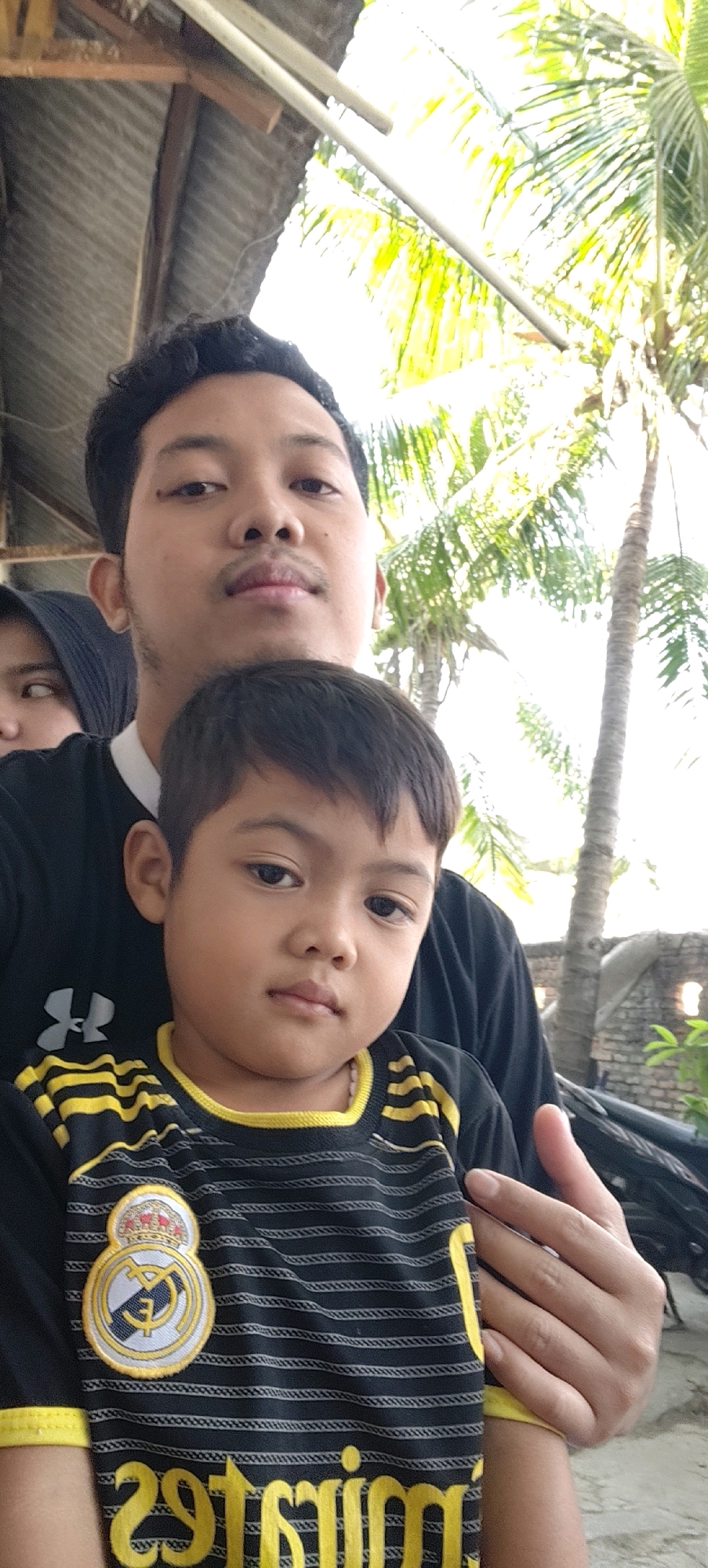 In Mekarmukti, Indonesia, a close-up, waist-up portrait captures a man holding a young boy in front of him. The man, seen from the chest up, is positioned behind the boy. He has dark, curly hair and a mustache. He is wearing a black t-shirt with a white collar. The boy is in the foreground, facing forward and looking towards the camera with a neutral expression. He has short, dark hair. He is wearing a black t-shirt with yellow stripes and a yellow collar. On the left side of his shirt, there is a crest with a circular emblem. Behind the subjects, palm trees and a rustic roof structure are visible against a bright, slightly overexposed sky, suggesting an outdoor setting. The overall lighting is bright, indicative of daylight.