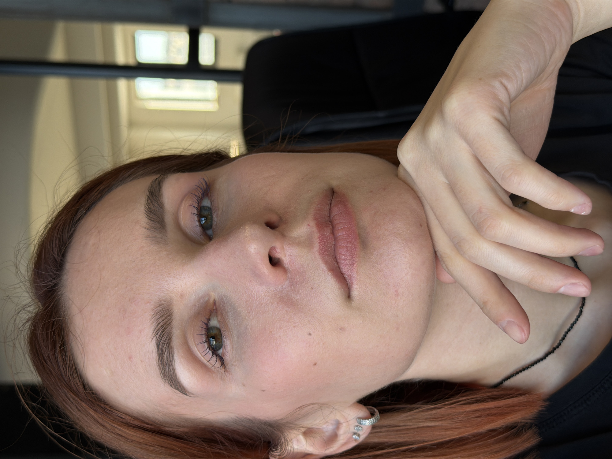 A close-up shot of a woman's face, tilted to the left. Her hand is resting on her chin. She has fair skin, light brown hair with reddish highlights, and green eyes. She is wearing a black shirt and a delicate black necklace. Her ear is pierced with multiple silver earrings. The background is blurry and includes a window with a view of a building. No information can be extracted regarding the location in the city of Chaikovsky, Russia.