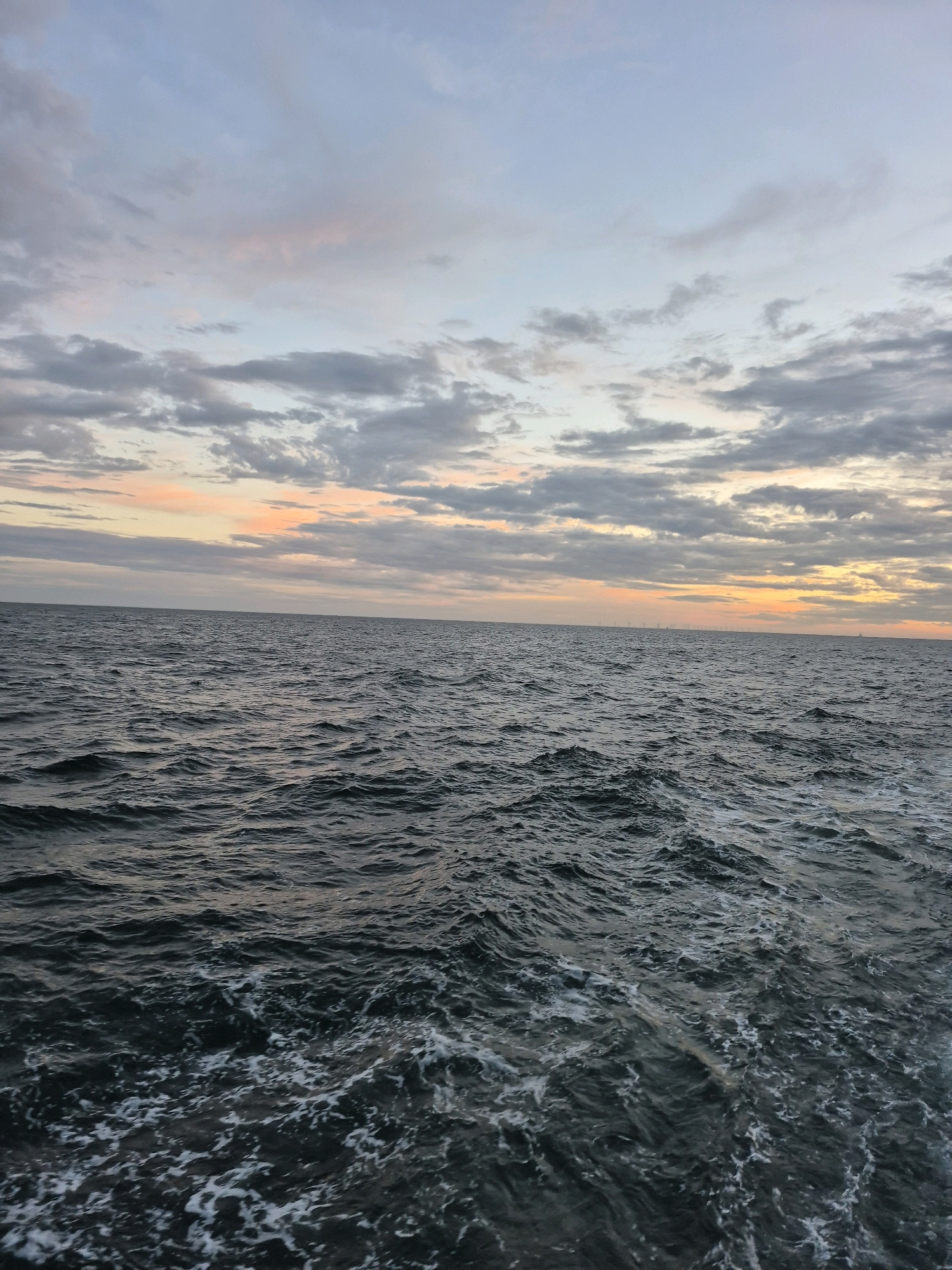 The image depicts a wide expanse of choppy, dark ocean water under a sky filled with soft, cloudy formations. The horizon is visible, marked by a pale band of light where the sky meets the sea. The clouds are varied in color, with shades of grey, white, and hints of pink and orange from what appears to be a sunset or sunrise. The water's surface is rippled, with white foam appearing in the foreground, suggesting movement and waves. In the far distance, along the horizon, a faint line of what appear to be structures, possibly windmills, can be discerned. The scene is peaceful and natural, with no human presence visible. The location is identified as Unknown, Germany.