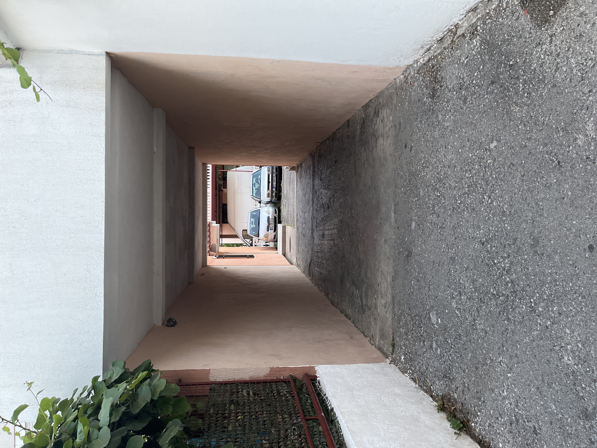 The image depicts a narrow alleyway leading towards two parked cars, a white van and a silver sedan. The alley has a rough asphalt surface and is flanked by peach-colored walls on both sides. The ceiling of the alley is a light gray concrete overhang. Overhanging greenery from the left side of the image is visible against a white building. The scene appears to be in a residential or commercial area in Budva, Montenegro, given the context provided. The lighting suggests it is daytime, possibly late afternoon. No people are visible in the frame.
