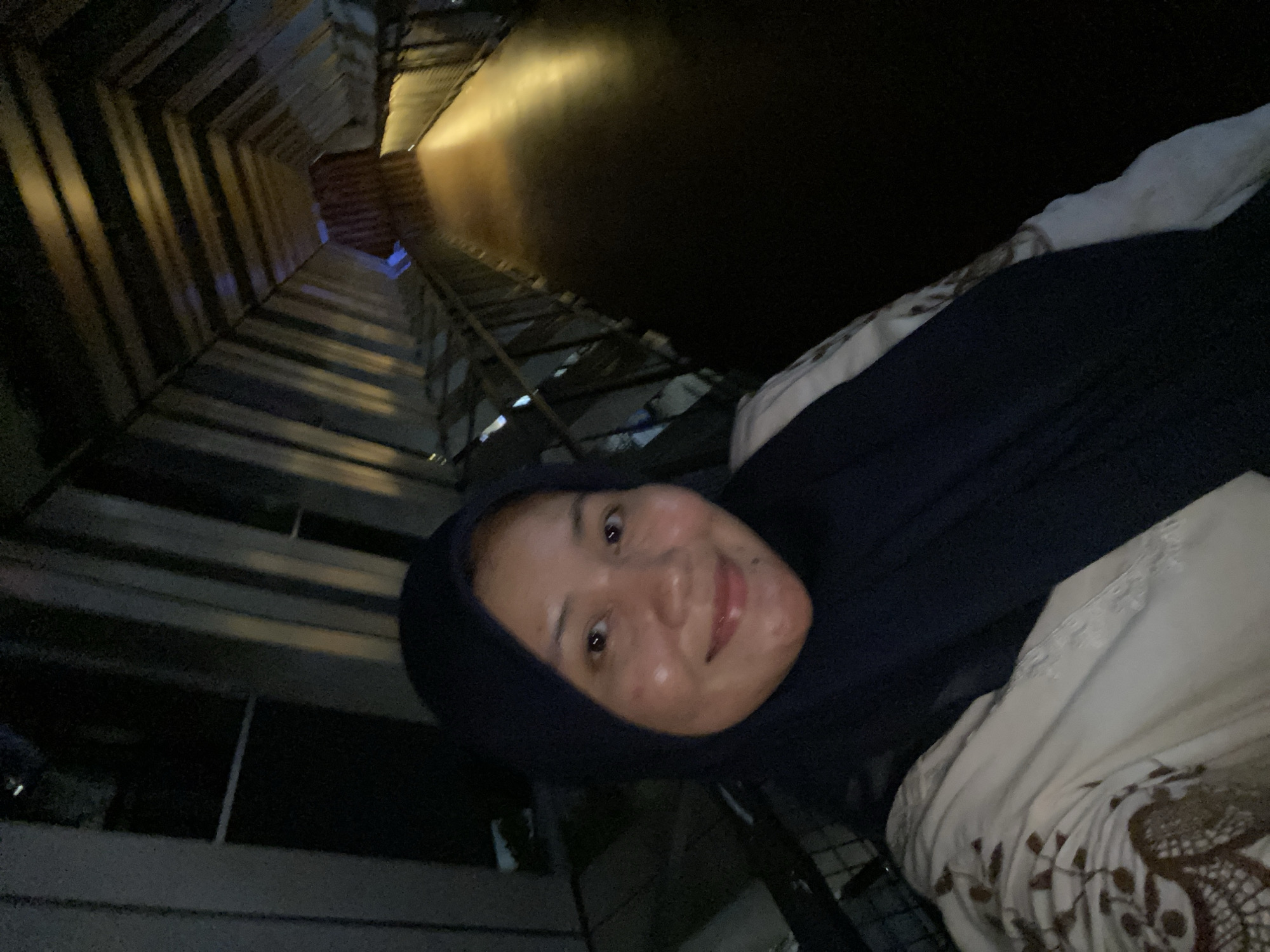 In Banjarbaru, Indonesia, a selfie shows a woman wearing a dark blue hijab and a light-colored patterned garment. She is positioned on a walkway with a distinctive tunnel-like structure overhead. The walkway appears to be made of dark material, possibly wood or a dark-colored floor, illuminated by warm, directional lighting that creates a dramatic effect. The tunnel structure above is composed of angled, metallic-like panels, creating a geometric pattern that recedes into the distance. The overall impression is one of an indoor or covered walkway with modern architectural features. The woman's face is the primary focus in the foreground, with the background providing context for her location.