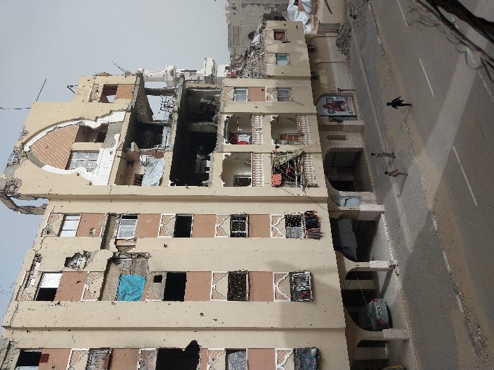 The image depicts a heavily damaged multi-story building in Khan Yunis, Palestinian Territory, overlooking a street. The building, approximately seven stories tall with a beige facade and reddish-brown decorative elements, exhibits extensive structural failure, particularly on its upper right side where large sections of walls and an arched architectural feature have collapsed. Numerous impact holes and pockmarks are visible across the entire facade. Windows are missing panes, shattered, or covered with blue tarps. Exposed rebar, concrete rubble, and charred interior sections are visible where walls have collapsed. Laundry is hanging from several windows and damaged balconies. The ground floor features arched open spaces; a silver sedan is parked under one on the left.
On the street and sidewalk in front of the building, three individuals, appearing to be children, are present. Two are standing on the paved sidewalk: one in light clothing closer to the building, and another in darker clothing slightly further out. The third individual, wearing dark clothing, is on the asphalt street, running or walking quickly away from the other two. The sky is a uniform light gray, indicating overcast conditions or haze. In the distance to the right, other structures with visible damage or white tarps are partially discernible. Overhead wires are visible at the top and bottom right of the frame.