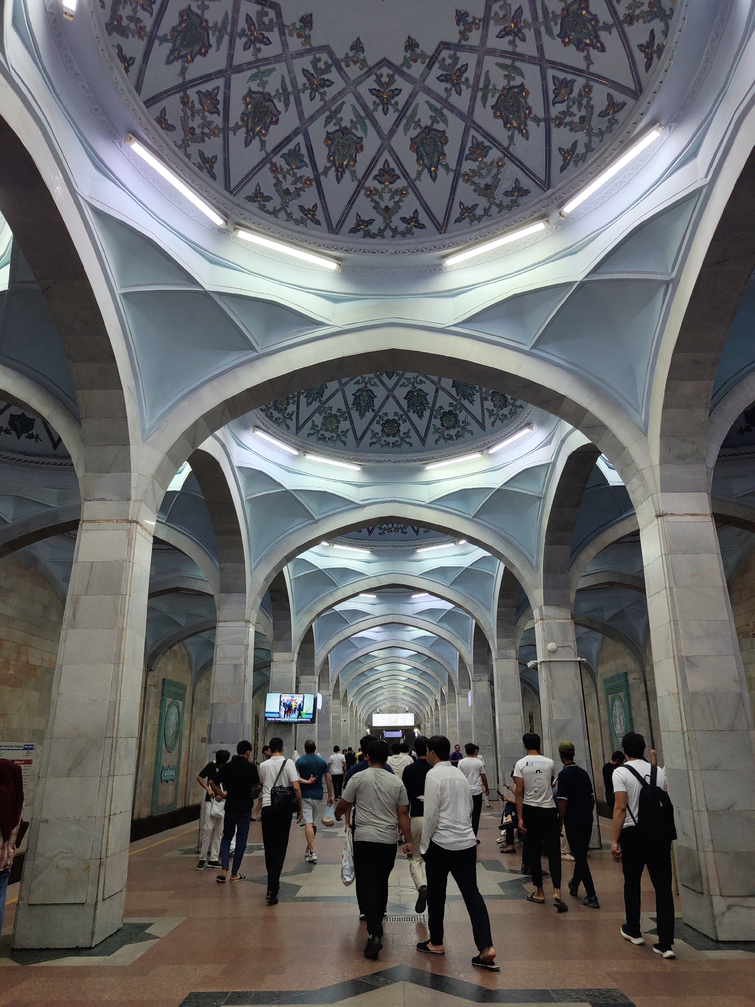 The image captures the interior of the Kosmonavtlar Metro Station in Tashkent, Uzbekistan. The station features a grand, cavernous space with a high, domed ceiling adorned with intricate geometric and floral patterns in blue and gold. The ceiling is supported by rows of large, marble-clad pillars that form a series of arches, creating a repetitive, receding perspective. Fluorescent lights are embedded in the ceiling.
A platform runs along the side of the station, where a group of people are walking. The individuals, dressed in casual attire, are moving in various directions, some walking towards the camera and others away from it. They are predominantly young men, with a few women visible. The flooring of the platform is a mix of patterned tiles. A large screen displaying a news program is mounted on the wall in the background. The overall impression is one of an architecturally significant and well-trafficked public space.