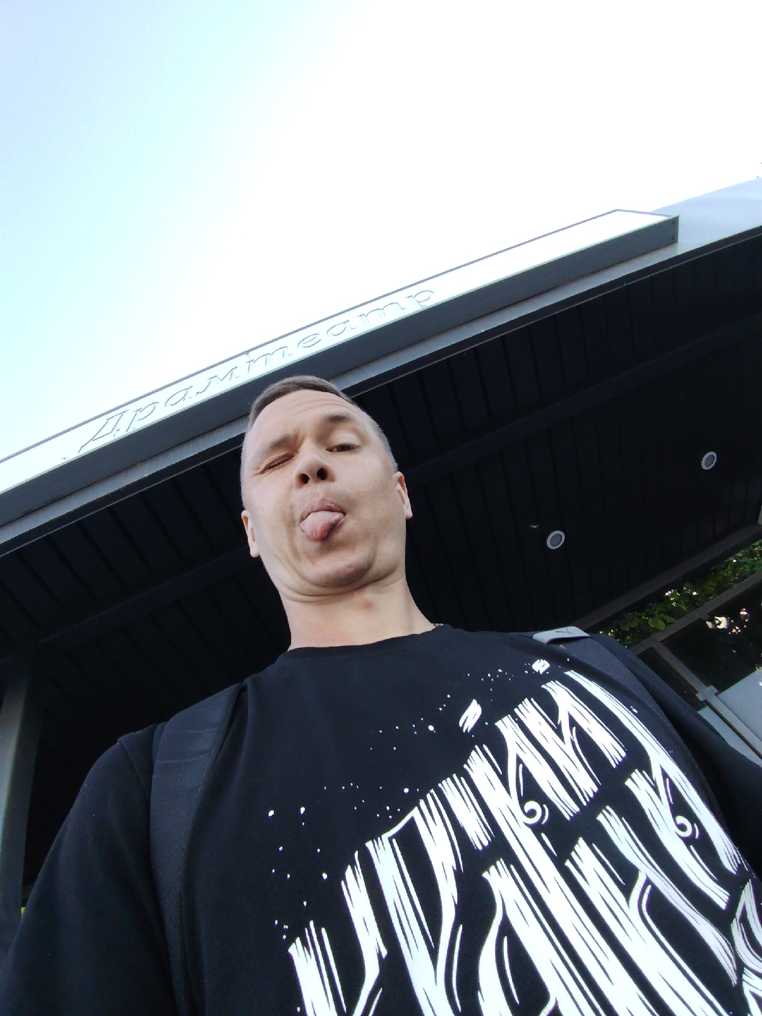 The photograph shows a man standing in front of the Drama Theater in the city of Chaikovsky, Russia. He is fair-skinned with short-cropped hair, wearing a black t-shirt with a white graphic design, and a backpack. The man is sticking out his tongue and winking. The building above and behind the man has dark horizontal planks, with recessed round lights. The name of the theater is displayed in Cyrillic. The sky is visible in the upper left corner.