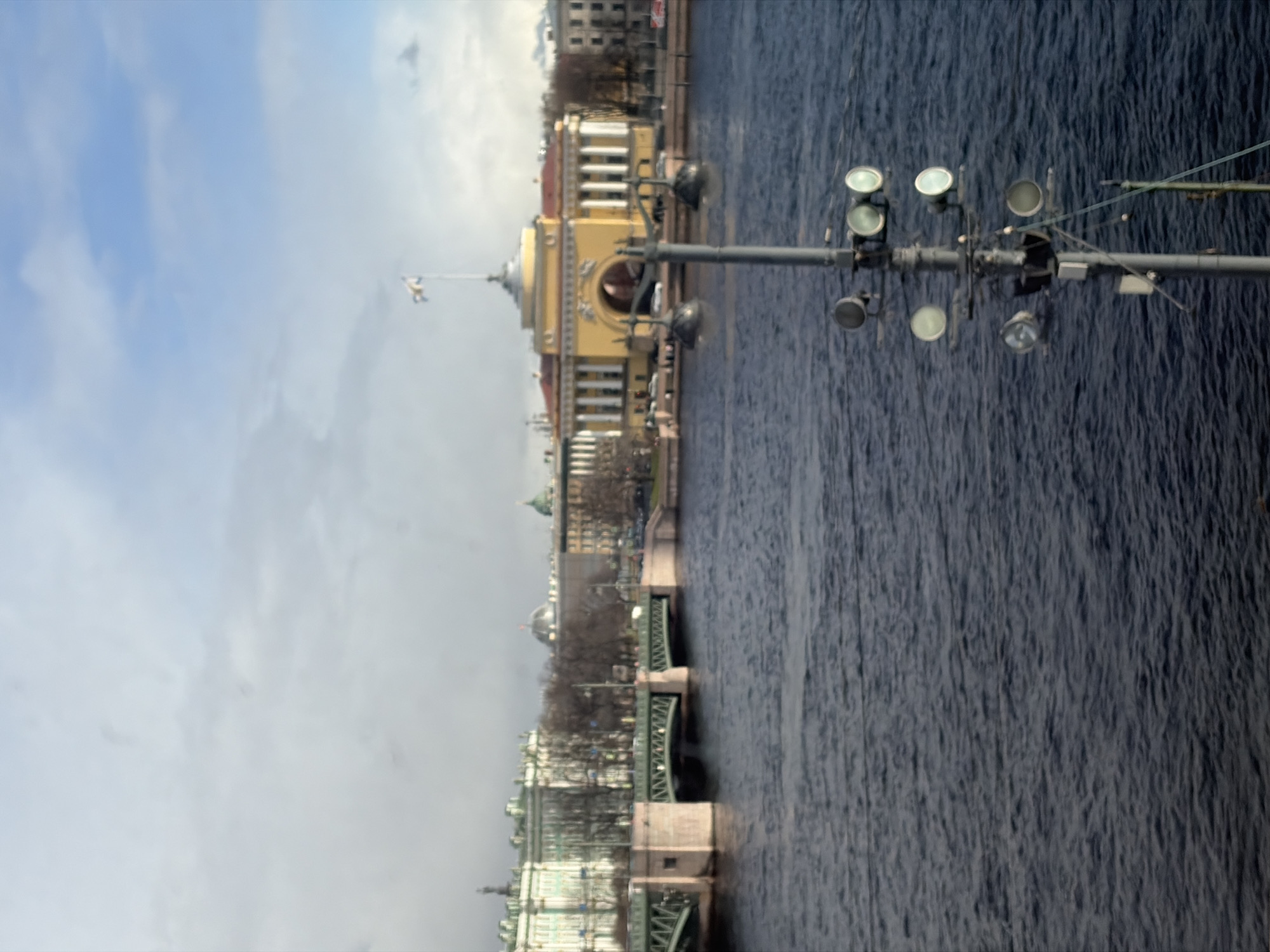 The image depicts a view in Saint Petersburg, Russia, showing a river, likely the Neva. A bridge spans the river, and a yellow building with a dome and spires is visible along the riverbank. Streetlights are positioned on the bridge. The sky is overcast, and the river appears dark with ripples.