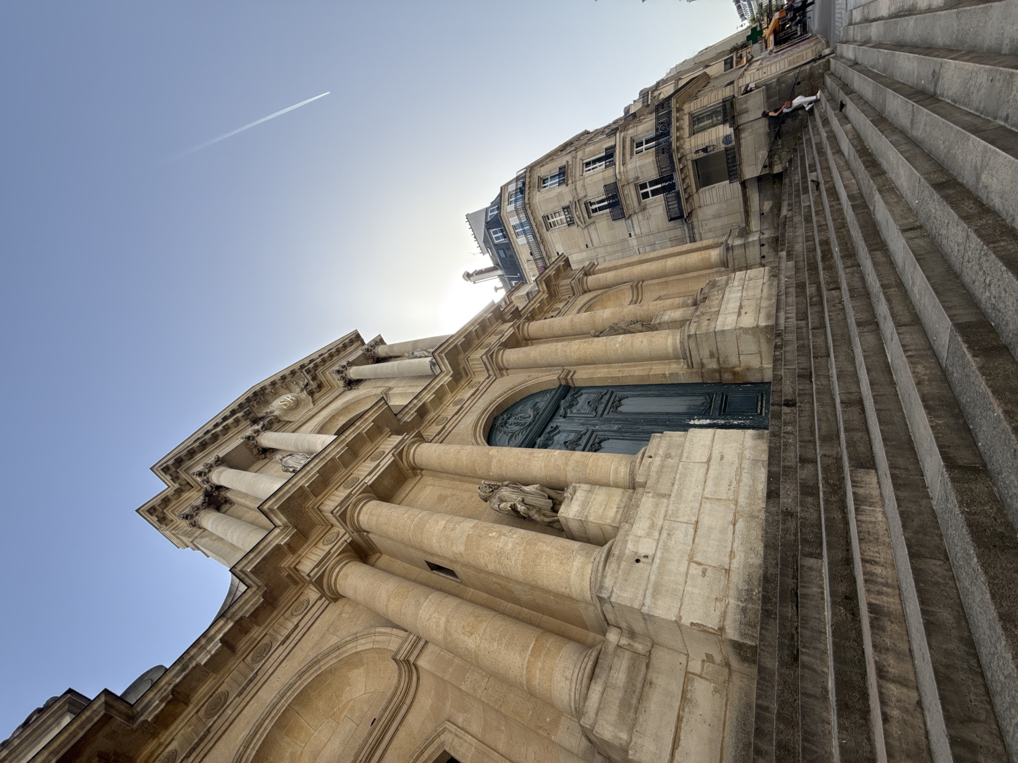 The image shows a low-angle view of the Église Saint-Sulpice in Paris, France. The church's façade, constructed of light-colored stone, is prominently featured, with classical architectural elements such as columns, pilasters, and a large arched doorway with a dark-colored door. There are several statues on the exterior of the building. Adjacent to the church, a wide set of stone steps lead upwards towards another building with apartments. In the upper portion of the frame, a few people can be seen at the top of the stairs. A contrail streaks across the clear blue sky above the church.