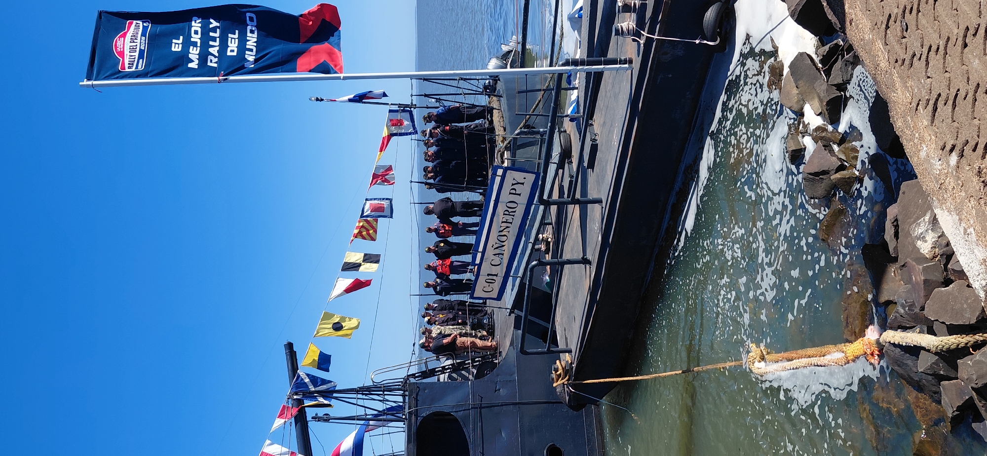 The image depicts a clear day in Encarnación, Paraguay, likely at the port or a waterfront area. In the foreground, a large, dark-colored vessel, identified by signage as "C-01 CANONERO PY," is docked. The vessel appears to be a patrol boat or a small warship, with a prominent gun mounted on its deck. A rope connects the vessel to the shore, which is lined with large, dark rocks.
A group of individuals, predominantly men in uniform and some in civilian attire, are gathered on the deck of the "C-01 CANONERO PY." Some appear to be military personnel, possibly navy officers or crew, given their uniforms. Others are in casual clothing, suggesting they might be guests, organizers, or spectators. The gathering seems formal, with people standing attentively.
Decorating the ship and the surrounding area are numerous flags. A large blue and red flag with white lettering is prominently displayed on a pole to the right, advertising "RALLY DEL PARAGUAY" and "EL MEJOR RALLY DEL MUNDO" (The Best Rally in the World). A string of international maritime signal flags is strung across the deck of the vessel, adding a nautical and celebratory element to the scene.
The background shows a calm body of water, likely the Paraná River, under a bright blue sky. The overall atmosphere suggests an event or ceremony taking place, possibly related to a rally or a naval demonstration. The presence of the "C-01 CANONERO PY" and the international flags points to a significant maritime or sporting event in Encarnación.
