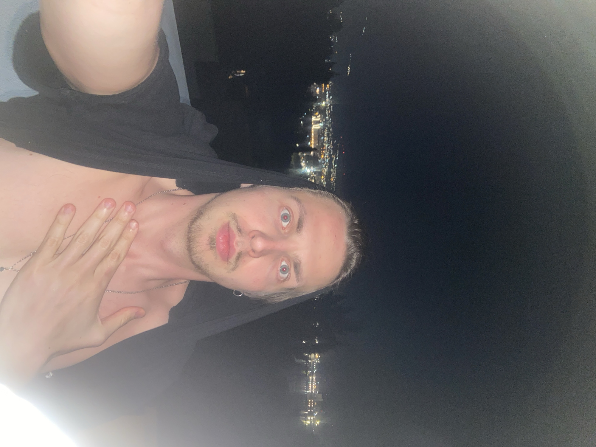 A selfie taken at night depicts a light-skinned male in Šušanj, Montenegro, against a blurred cityscape background. The man is partially unclothed, wearing a black garment around his shoulders and chest, with a chain necklace visible around his neck. His left hand rests on his upper chest. The background shows the dark night sky and blurred city lights of Šušanj.