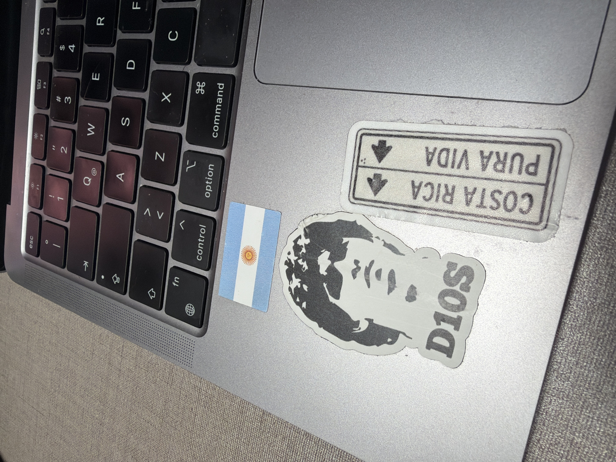The image shows a close-up view of a silver laptop keyboard with several stickers on its surface. The stickers include the flag of Argentina, a black and white portrait of a person with the text "D10S" below it, and a rectangular sticker with the text "COSTA RICA PURA VIDA" and arrows pointing in opposite directions. The laptop is placed on a textured surface that appears to be a table or desk. There are no people or explicit signs of human presence in the immediate scene. The lighting suggests it is indoors, possibly in a hotel room or a workspace. The location is not discernible from the image itself; information about its placement in Cancún, Mexico, is external to the visual content.