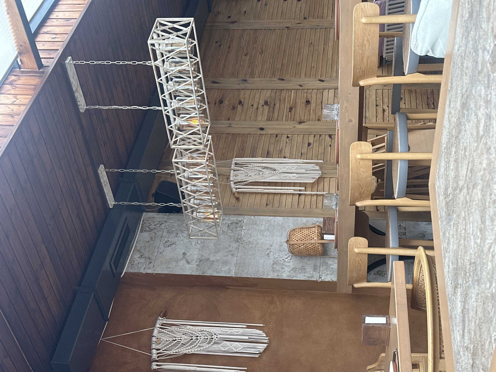 The image shows an indoor scene, likely a restaurant or dining area in Tsakhkadzor, Armenia, recognizable by the wooden architecture style typical of the region. The viewpoint is looking up towards the ceiling. A decorative, geometric chandelier hangs from the wooden ceiling. A second, similar but smaller chandelier is also present. Below the light fixtures, a woven basket is seen on the floor, possibly for decorative or storage purposes.
To the right of the basket are wooden dining chairs and tables, indicating a seating area. The walls are adorned with macrame wall hangings, adding a bohemian touch to the space. The floor appears to be tiled with neutral colors. The building appears to be made primarily of wood.