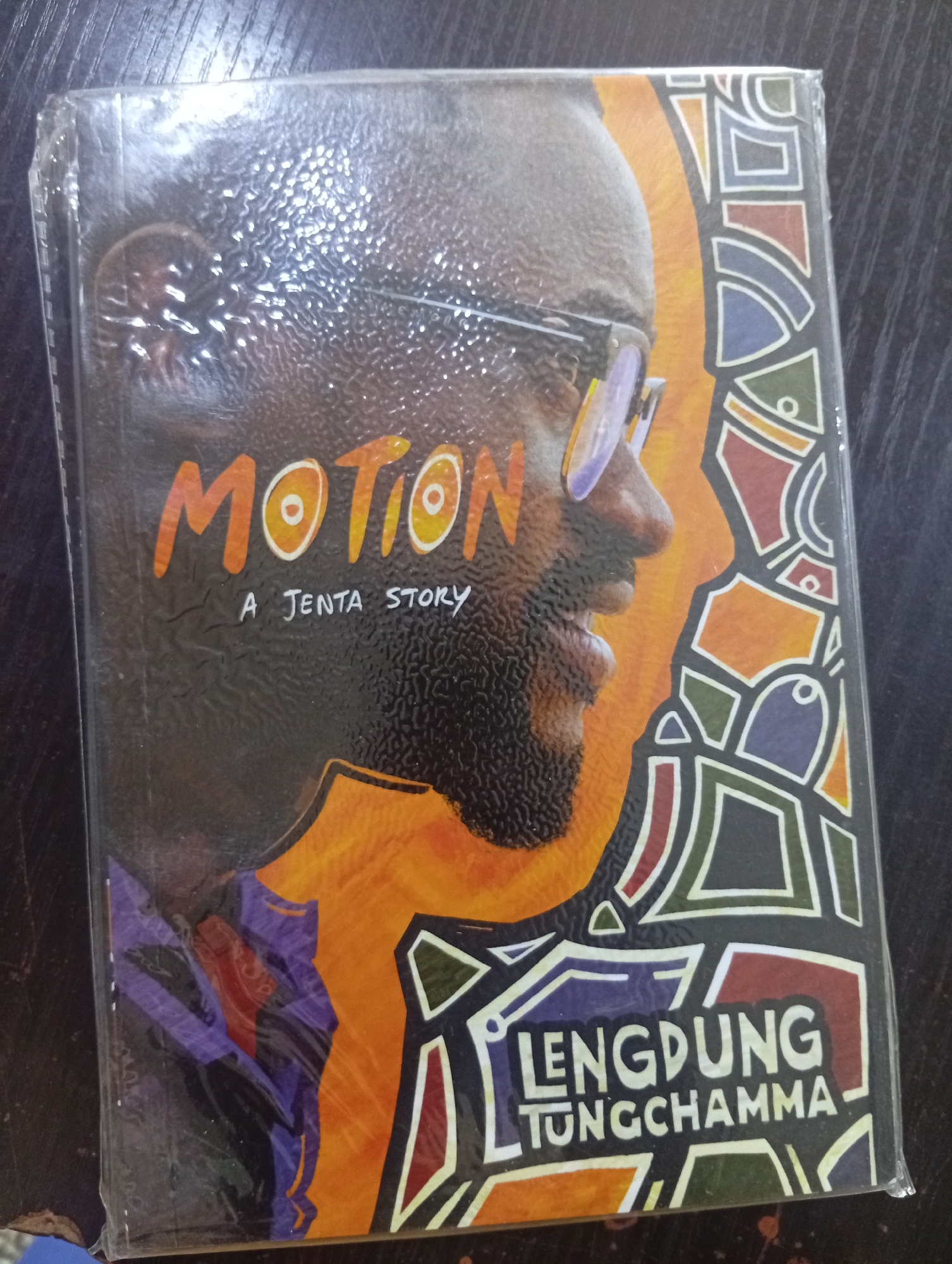 The image shows the cover of a book titled "MOTION A JENTA STORY". The cover features a close-up profile of a Black man wearing glasses, with an orange and black abstract design behind him. The book is shrink-wrapped and placed on a dark, textured surface, likely a table or desk. The lighting is overhead, creating reflections on the plastic wrap. No specific location in Jos, Nigeria is identifiable from the image itself.