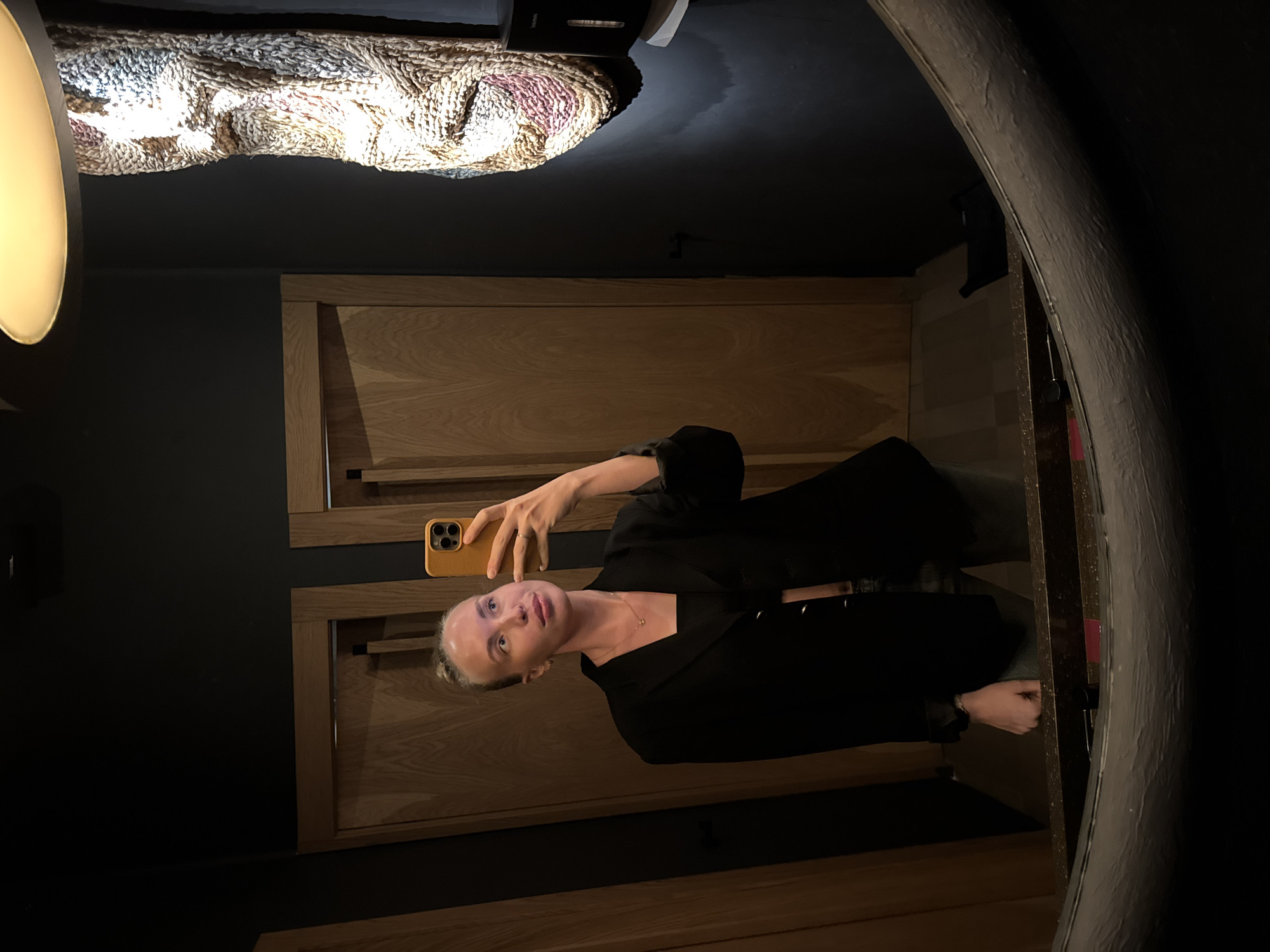 A woman is taking a selfie in a mirror. She is wearing a black blazer. The mirror is in a dark room with wooden panels on the walls. There is a decorative light fixture on the ceiling. The location is in Sochi, Russia.