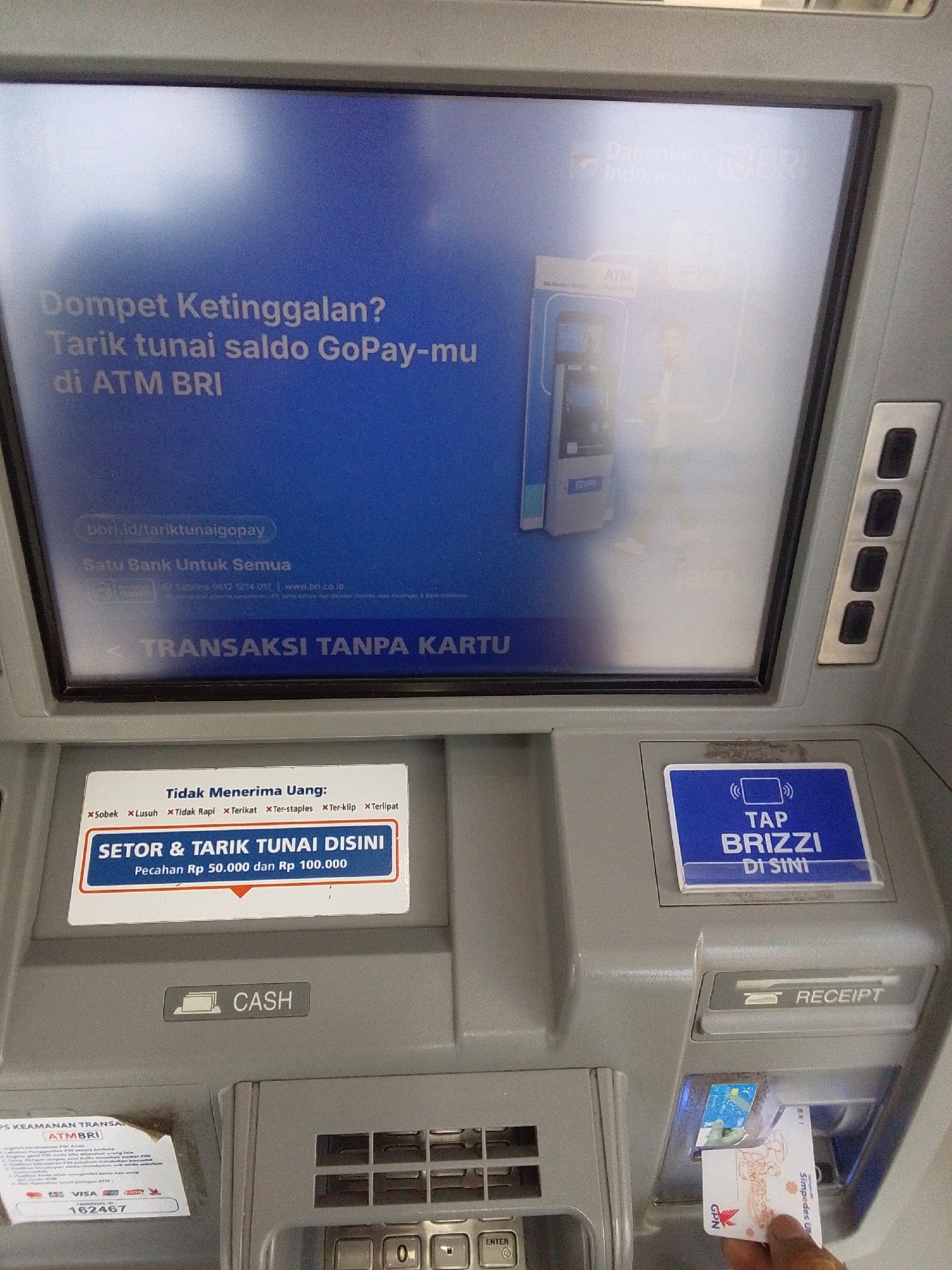 A close-up, eye-level, medium shot captures an ATM machine, possibly in Purworejo, Indonesia, with its screen displaying information and a card being inserted. The ATM screen, illuminated and taking up the top two-thirds of the frame, features a blue background with white text and graphics. The text in white says "Dompet Ketinggalan? Tarik tunai saldo GoPay-mu di ATM BRI". Below this, in smaller text, is "bbri.id/tariktunaigopay" and "Satu Bank Untuk Semua". Further down, a line of text reads "< TRANSAKSI TANPA KARTU". The screen also shows an image of an ATM and a person.
Below the screen, a white label with red text lists items not accepted, such as "Sobek", "Lusun", "Tidak Rapi", "Terikat", "Ter-staples", "Ter-klip", and "Terlipat". Below this, a larger blue and white label reads "SETOR & TARIK TUNAI DISINI" with the text "Pecahan Rp 50.000 dan Rp 100.000".
To the right of this label, a blue sign displays white text that says "TAP BRIZZI DI SINI".
Further down, a grey slot is labeled "CASH". To the right, a slot is labeled "RECEIPT".
In the bottom right corner, a hand is inserting a white and blue card into a slot. The card has graphics and text, including "GPN" and "Simpedes UMKM BRI".
The overall environment appears to be an indoor public space, typical of a bank or ATM vestibule. The lighting is adequate to illuminate the ATM's screen and the card insertion area. The ATM machine itself is made of grey plastic and metal.