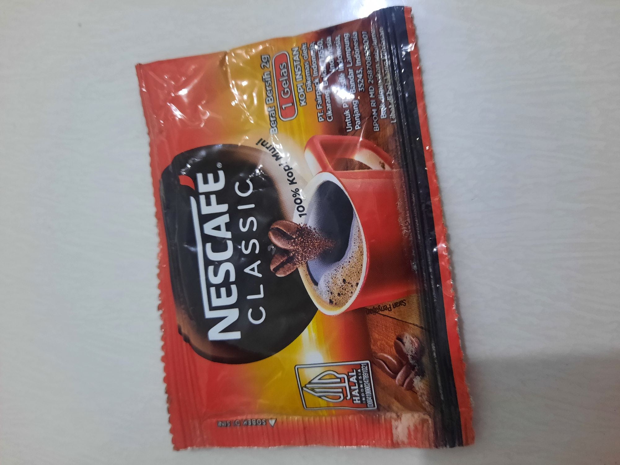 A Nescafé Classic instant coffee sachet is displayed against a white background. The sachet is predominantly red and black with yellow accents, featuring the Nescafé logo and text indicating "100% Kopi Murni" (100% Pure Coffee) and "1 Gelas" (1 Glass). A HALAL logo is visible on the left side of the packaging. The image includes a graphic of a red coffee cup with frothy coffee and coffee beans. There is no indication of the city of Lhokseumawe, Indonesia, within the image itself.