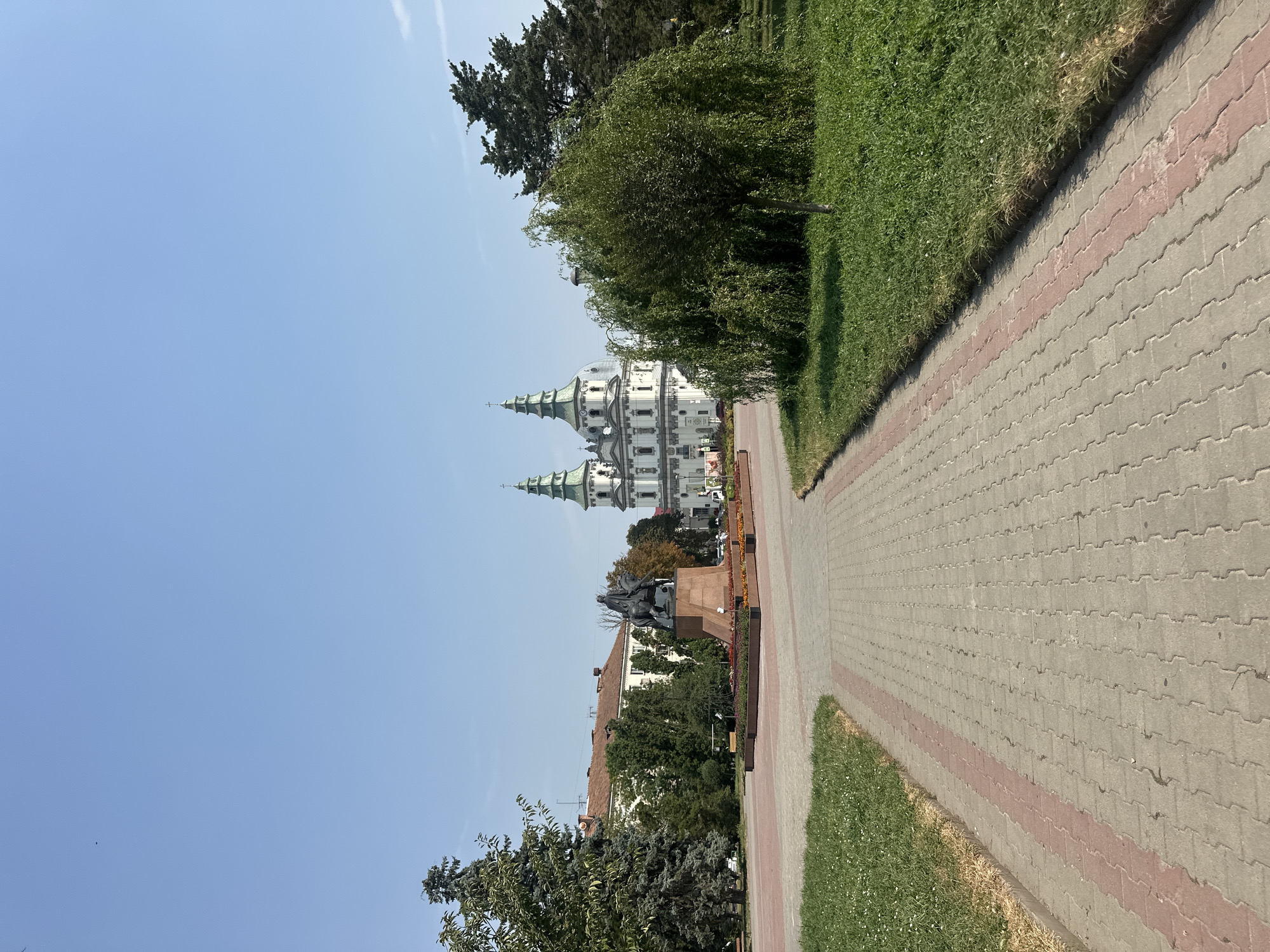 The image shows a wide, paved walkway leading towards a large, ornate white building with green spires, likely a church or cathedral. To the left of the walkway, there is a monument featuring a bronze equestrian statue on a stone pedestal. Beyond the monument, there are trees and buildings. To the right of the walkway, there is a grassy area with a large, leafy tree. The sky is clear and blue. The scene is located in Ternopil, Ukraine, specifically in front of the Ternopil Nativity Cathedral and near the monument to Ivan Franko.