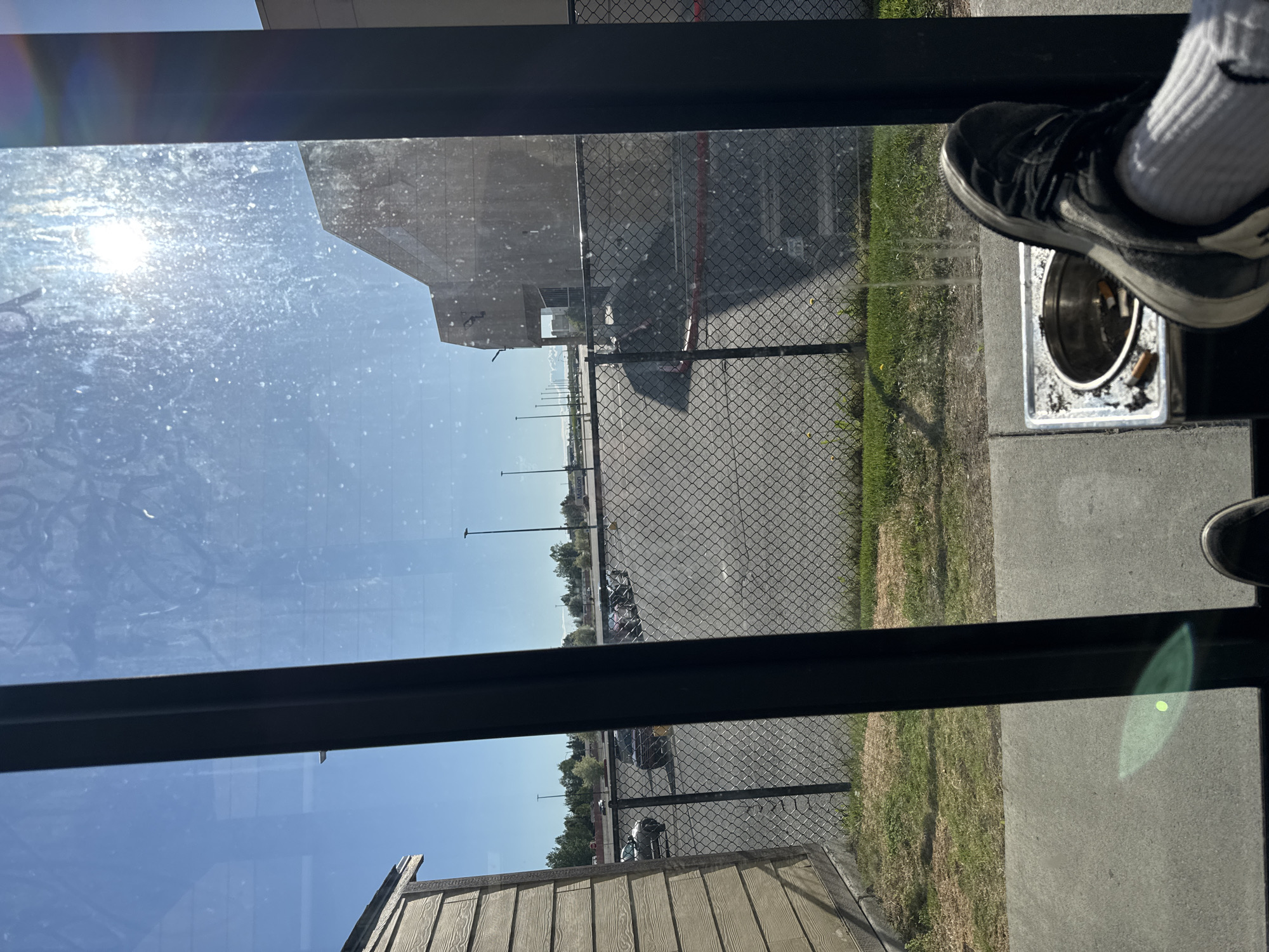 The image depicts a view from inside a structure, looking out through a glass pane that exhibits dirt and lens flare from the bright sun. The foreground shows a person's feet, one resting on a metal ashtray containing cigarette butts, and the other partially visible. The ashtray is positioned on a concrete surface. Beyond the glass, a chain-link fence encloses a paved area, likely a parking lot, with several cars parked in rows. A building, possibly a commercial establishment, is visible in the background, as are streetlights and trees. The sky is clear and blue. The location is the city of Anchorage, United States of America.