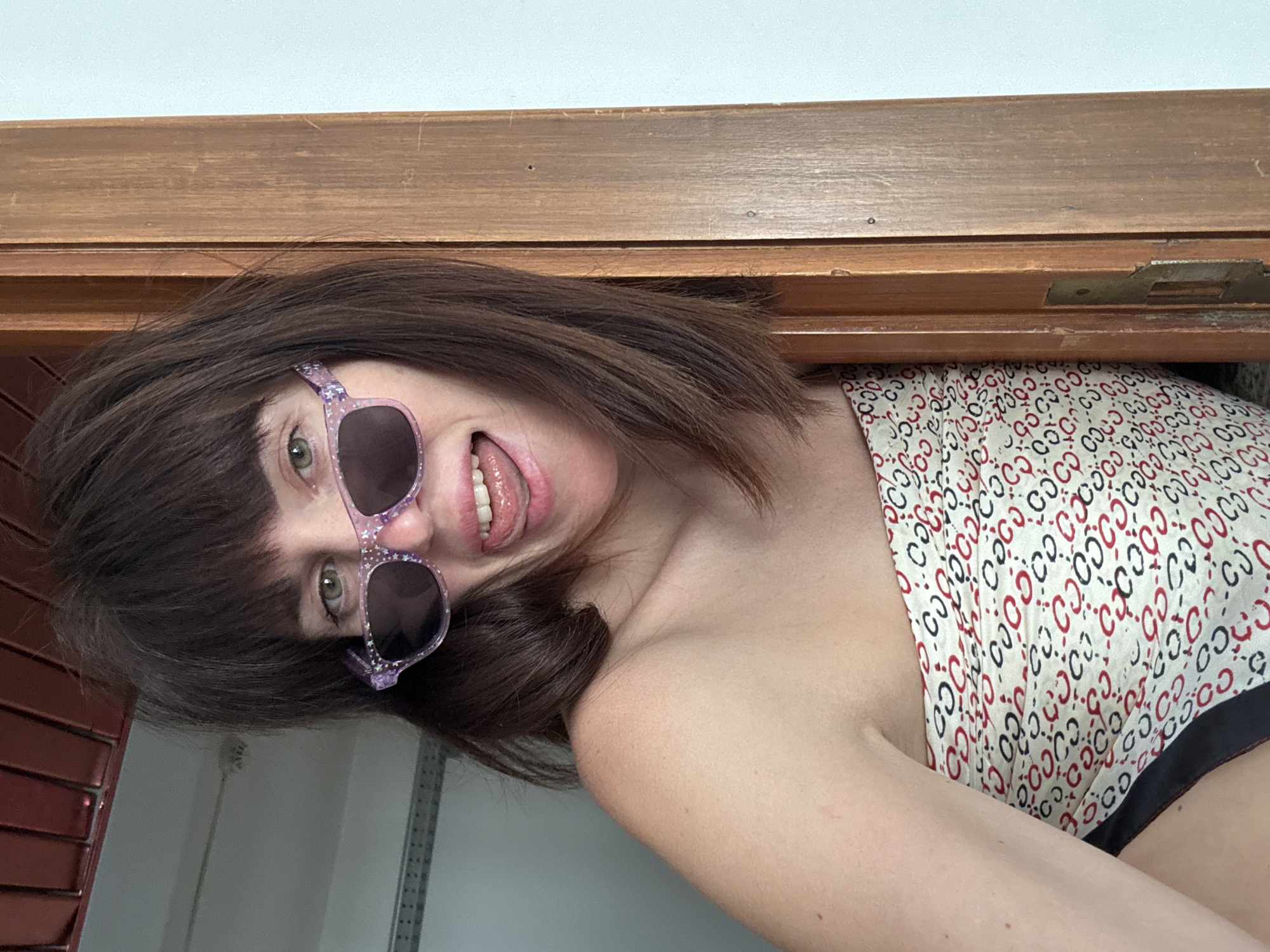 A medium shot shows a woman with brown hair and bangs, wearing purple sunglasses with white stars and a patterned strapless top. She is sticking her tongue out and leaning against a wooden door frame. The background consists of a gray wall with some metal shelving above and a white wall. The location is identified as Isola delle Femmine, Italy.