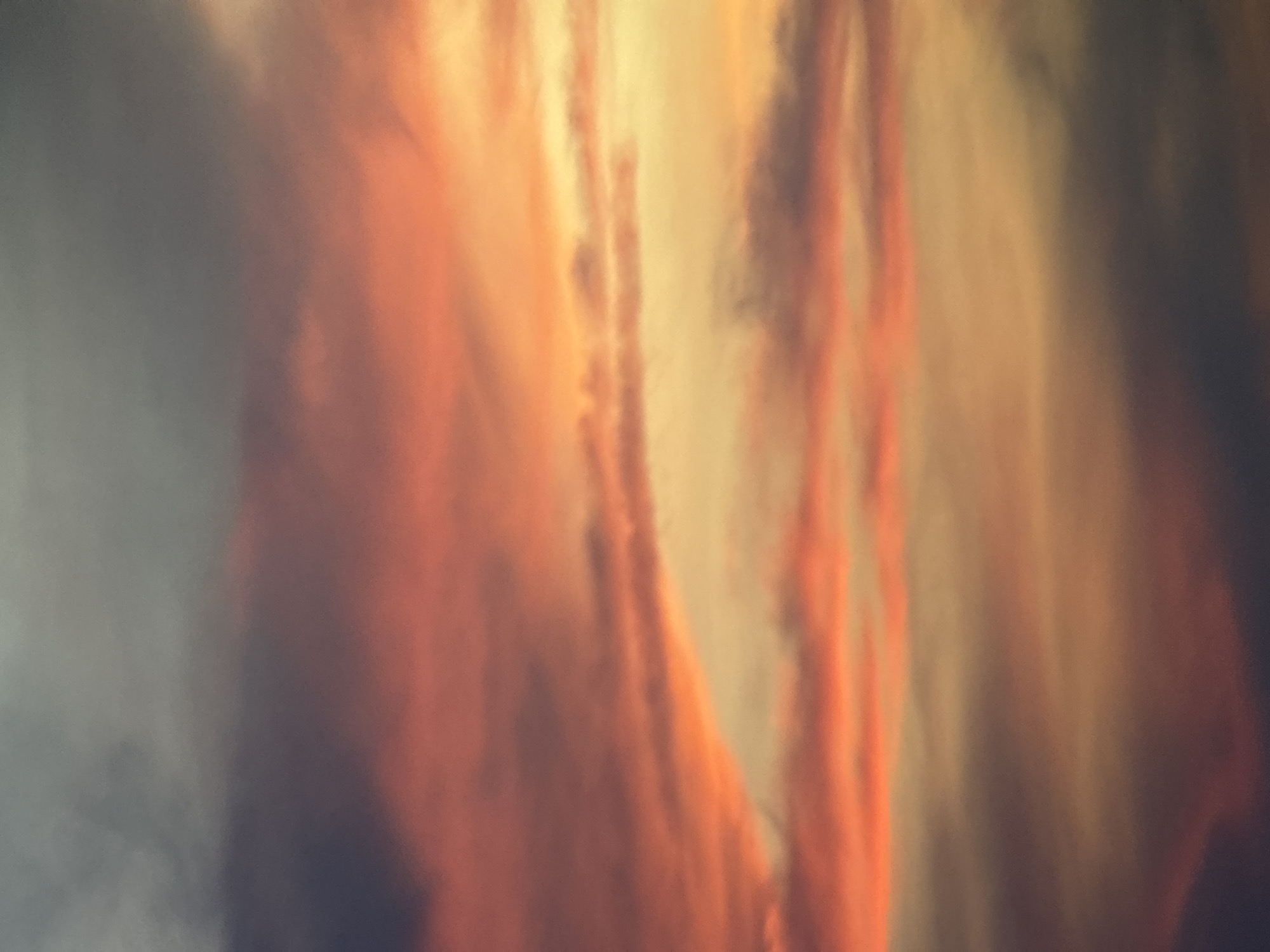 A vertical, slightly out-of-focus photograph captures a sky filled with clouds displaying the vibrant colors of a sunset over the city of Nha Trang, Vietnam. The clouds are arranged in horizontal bands, transitioning from dark gray at the top to shades of pink, orange, and yellow closer to the horizon. Some of the clouds appear wispy and thin, allowing the warmer hues of the setting sun to peek through. The overall impression is one of a dramatic and colorful sky, indicative of the natural beauty often seen in coastal Vietnamese cities during twilight. There are no people, subjects, objects, or actions depicted, as the image focuses solely on the atmospheric phenomenon.